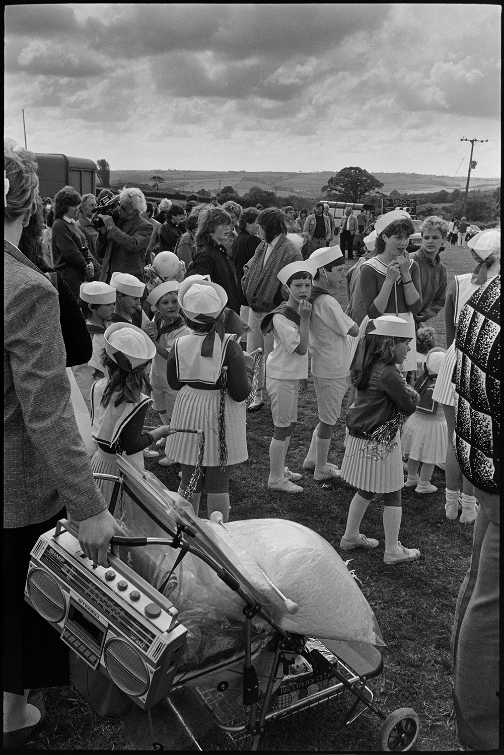 School fete, drum majorettes competition, early tractor display, mothers and children.
[People gathered in a field for the Chulmleigh School Fete. A group of majorettes are getting ready to perform. A woman with a pram and tape player is visible in the foreground.]