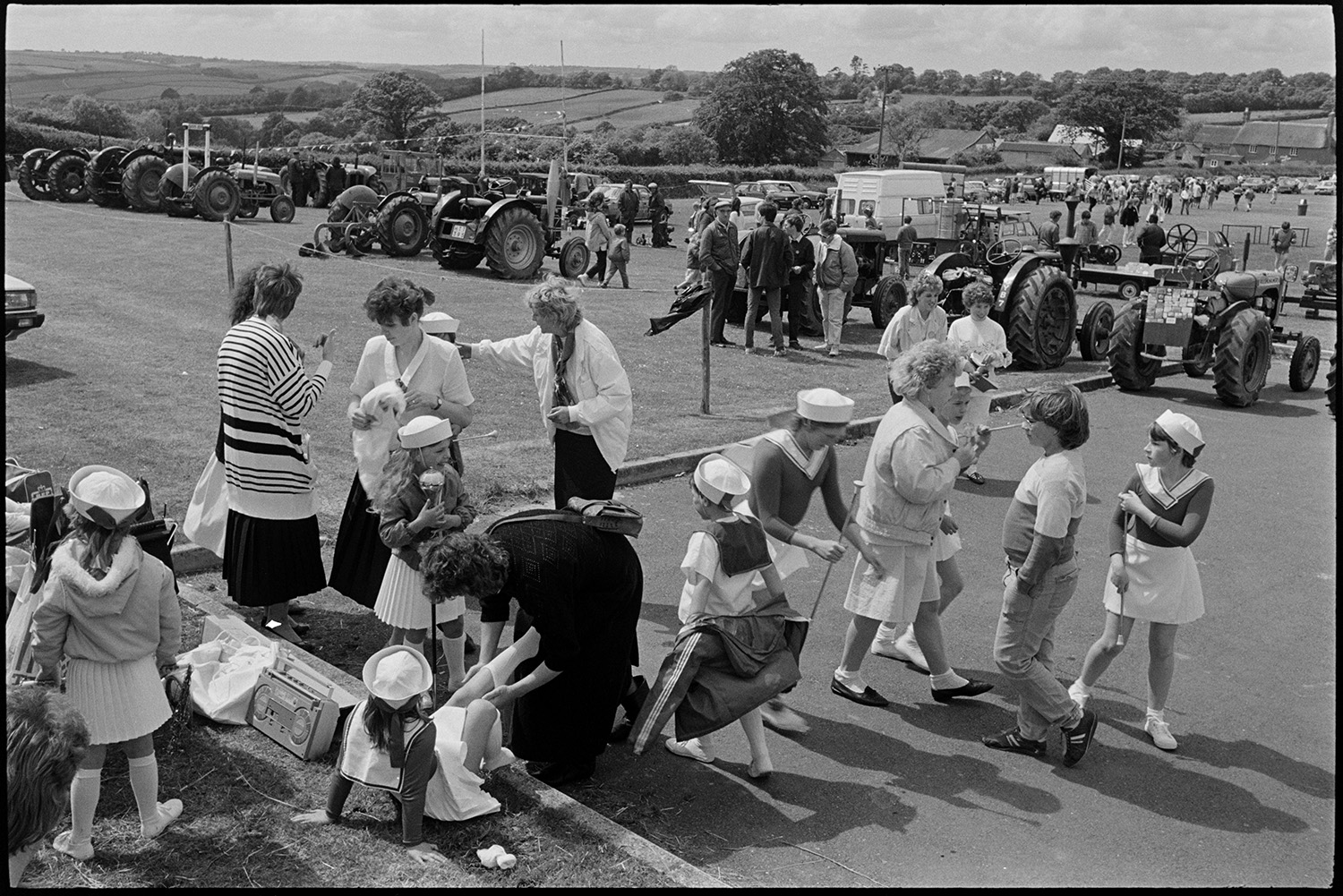 School fete, drum majorettes competition, early tractor display, mothers and children.
[People gathered in a field for the Chulmleigh School Fete. A group of majorettes are getting reading in the foreground and a display of vintage tractors is visible in the background.]