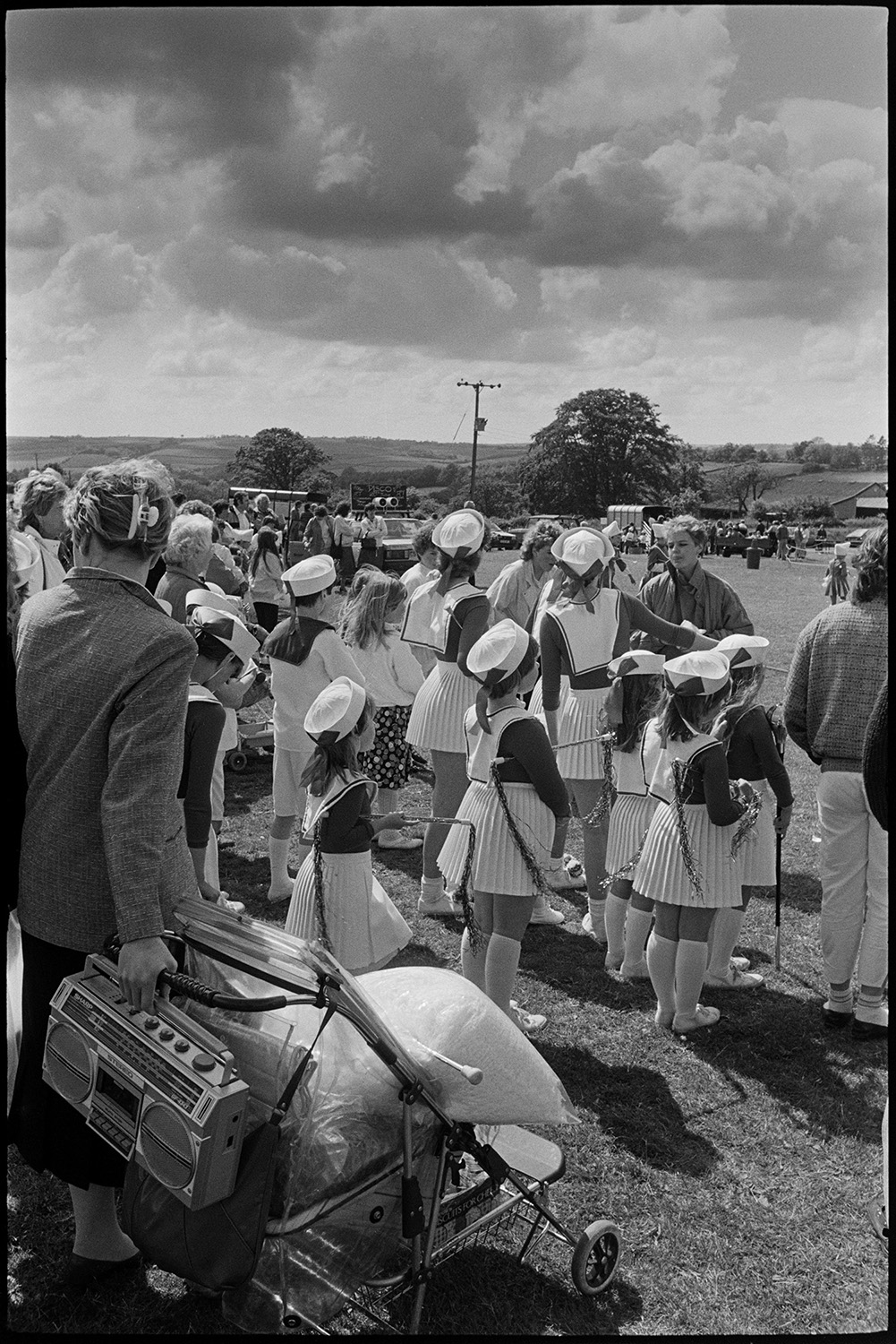 School fete, drum majorettes competition, early tractor display, mothers and children.
[People gathered in a field for the Chulmleigh School Fete. A group of majorettes are getting ready to perform. A woman with a pram and tape player is visible in the foreground.]