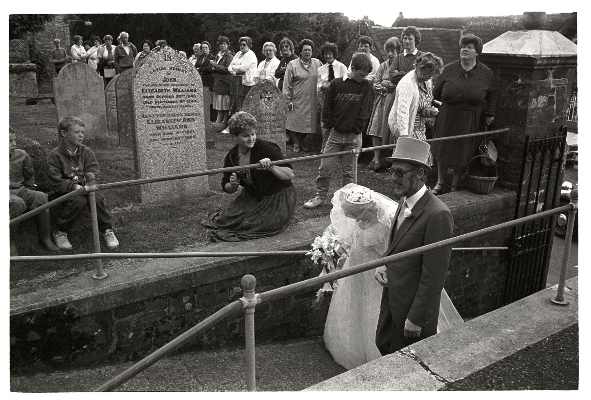 Onlookers at wedding, bride and father arriving in top hat. 
[Onlookers gathered in Chulmleigh churchyard to watch a bride and her father arrive for a wedding. The father of the bride is wearing a top hat.]