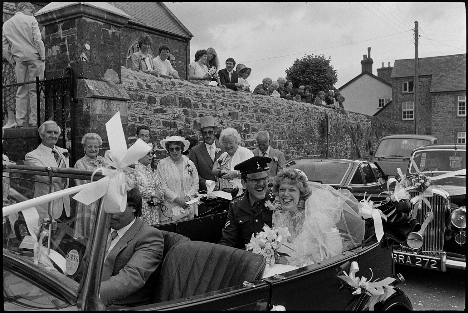 Onlookers at wedding, bride and groom arriving and leaving church in vintage car.
[A bride and groom, wearing a uniform, sitting in a vintage car after their wedding, with guests watching outside Chulmleigh Church.]