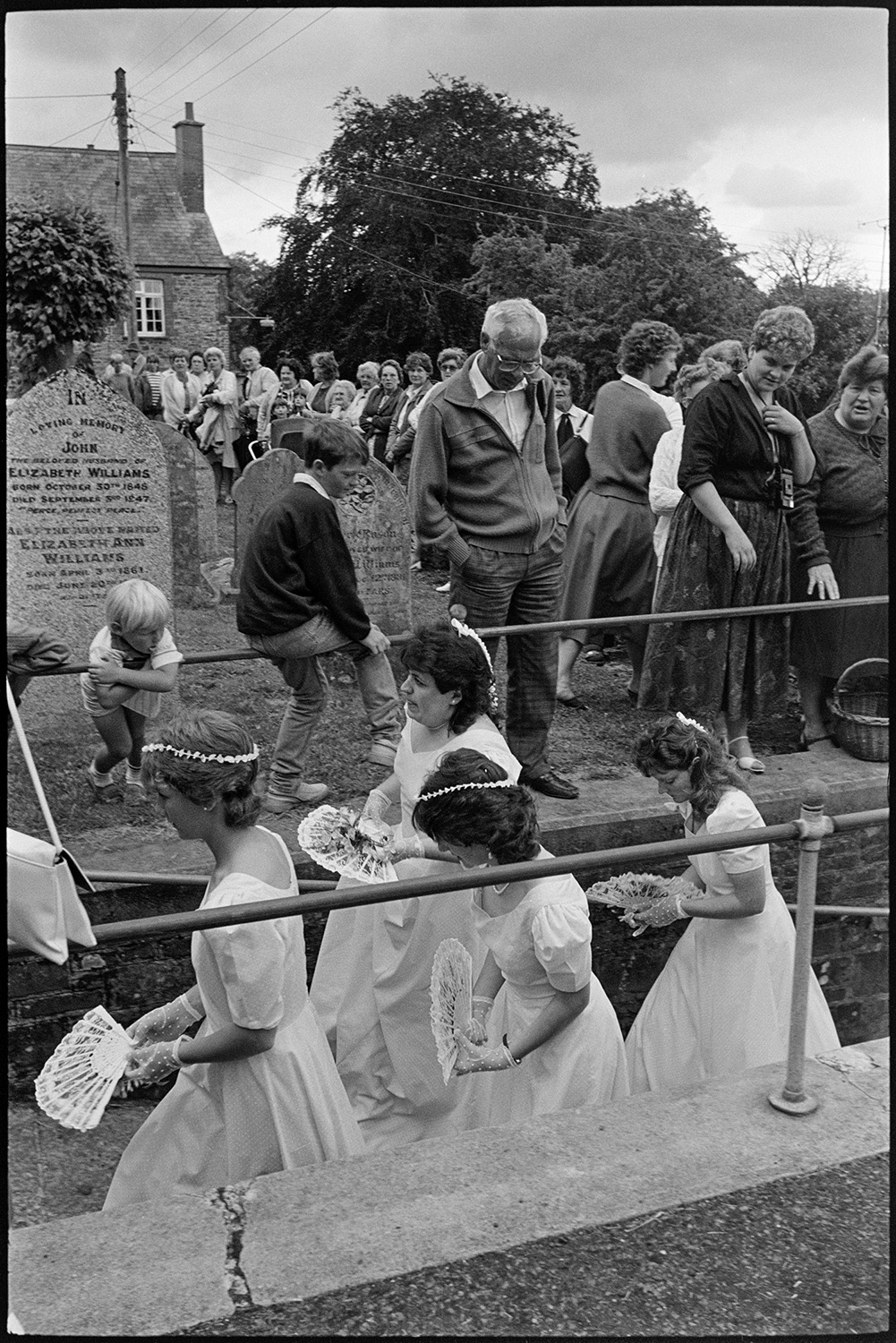 Onlookers at wedding, bride and groom arriving and leaving church in vintage car.
[Bridesmaids walking up Chulmleigh Church path to a wedding. Onlookers are gathered in the churchyard watching the procession.]