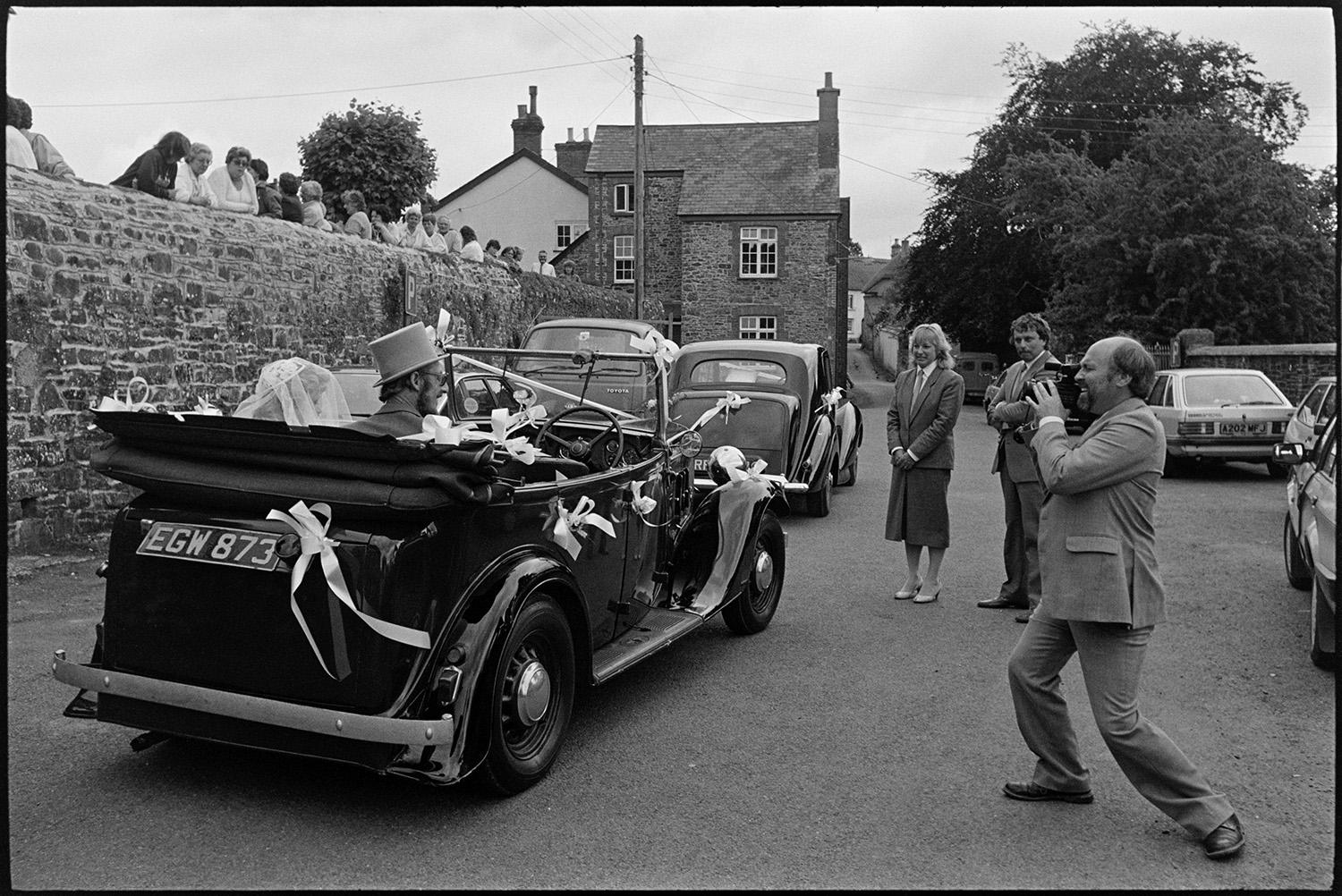 Onlookers at wedding, bride and groom arriving and leaving church in vintage car.
[A bride with her father arriving for her wedding at Chulmleigh Church in a vintage car. A man is videoing them and people are looking over the church wall. ]