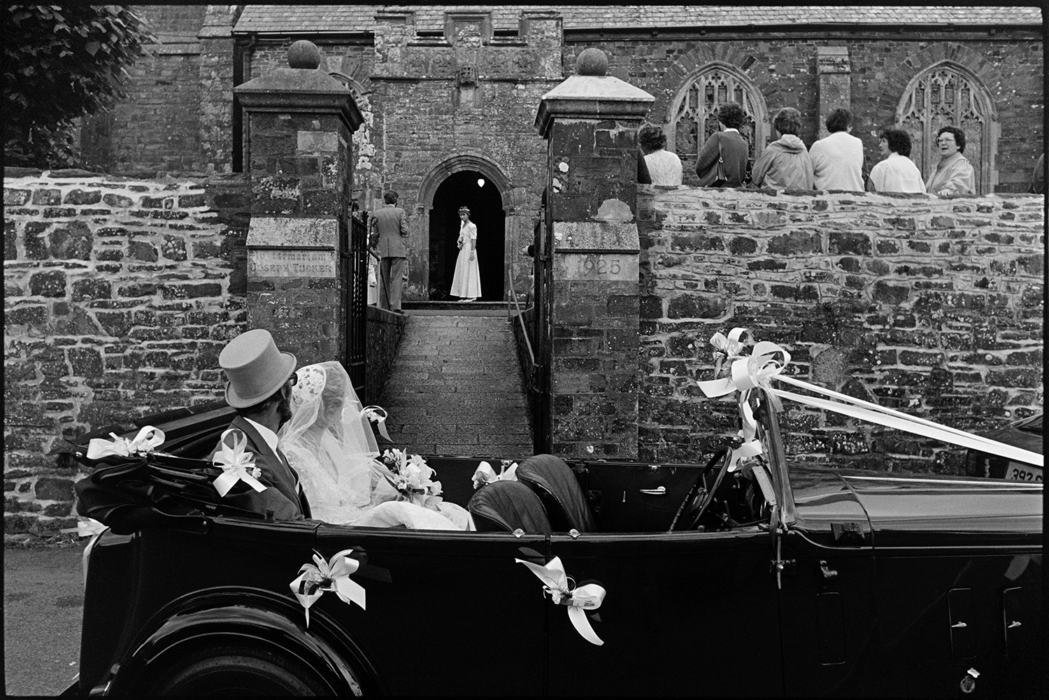 Onlookers at wedding, bride and groom arriving and leaving church in vintage car.
[A bride with her father arriving for her wedding at Chulmleigh Church in a vintage car decorated with ribbons. People are leaning on the church wall watching and a bridesmaid can be seen standing in the church porch.]