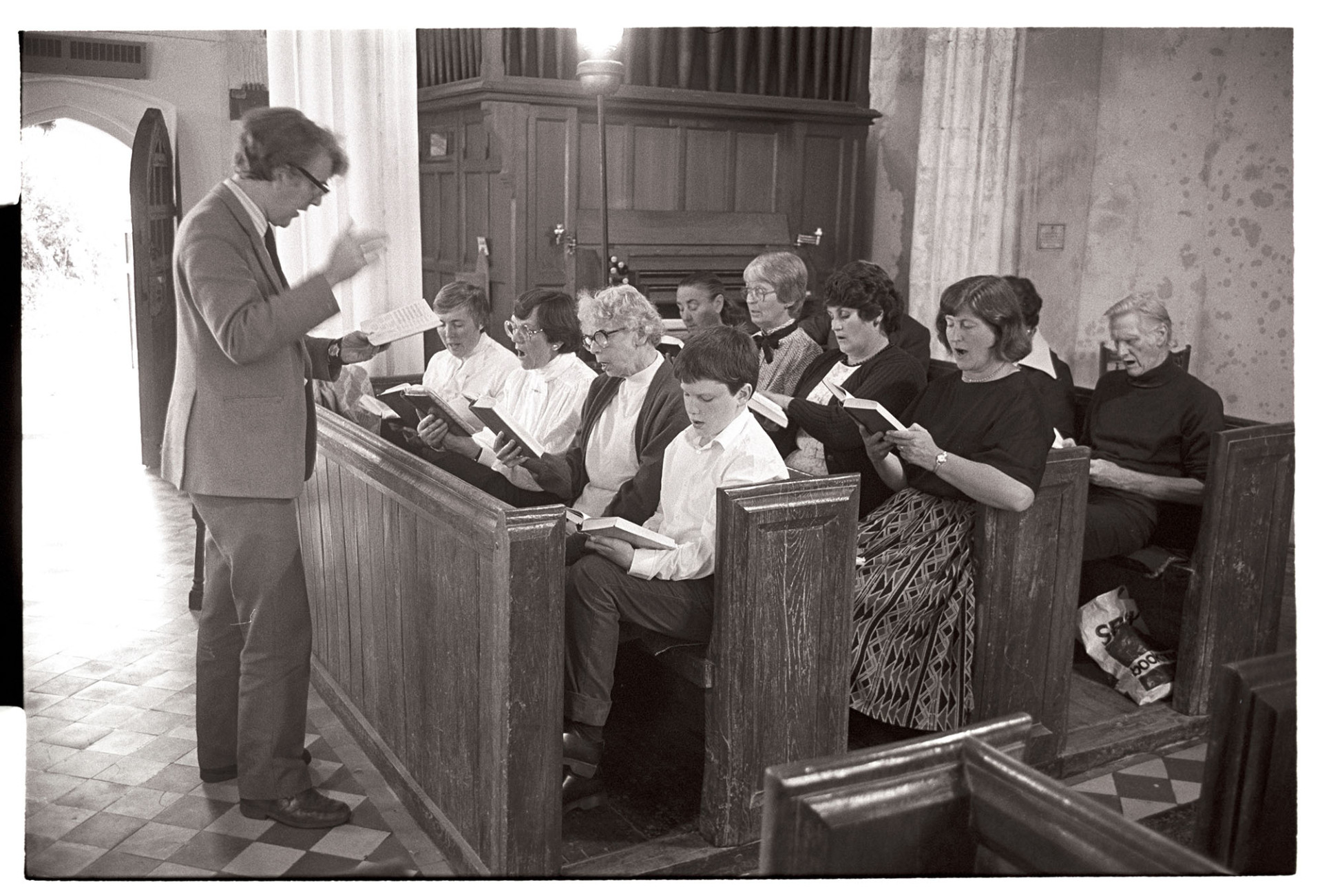 Choir practising in church. 
[David Davis conducting and singing with a choir who are practising at Warkleigh Church. They are sat in pews by the organ and the open church door is visible in the background.]