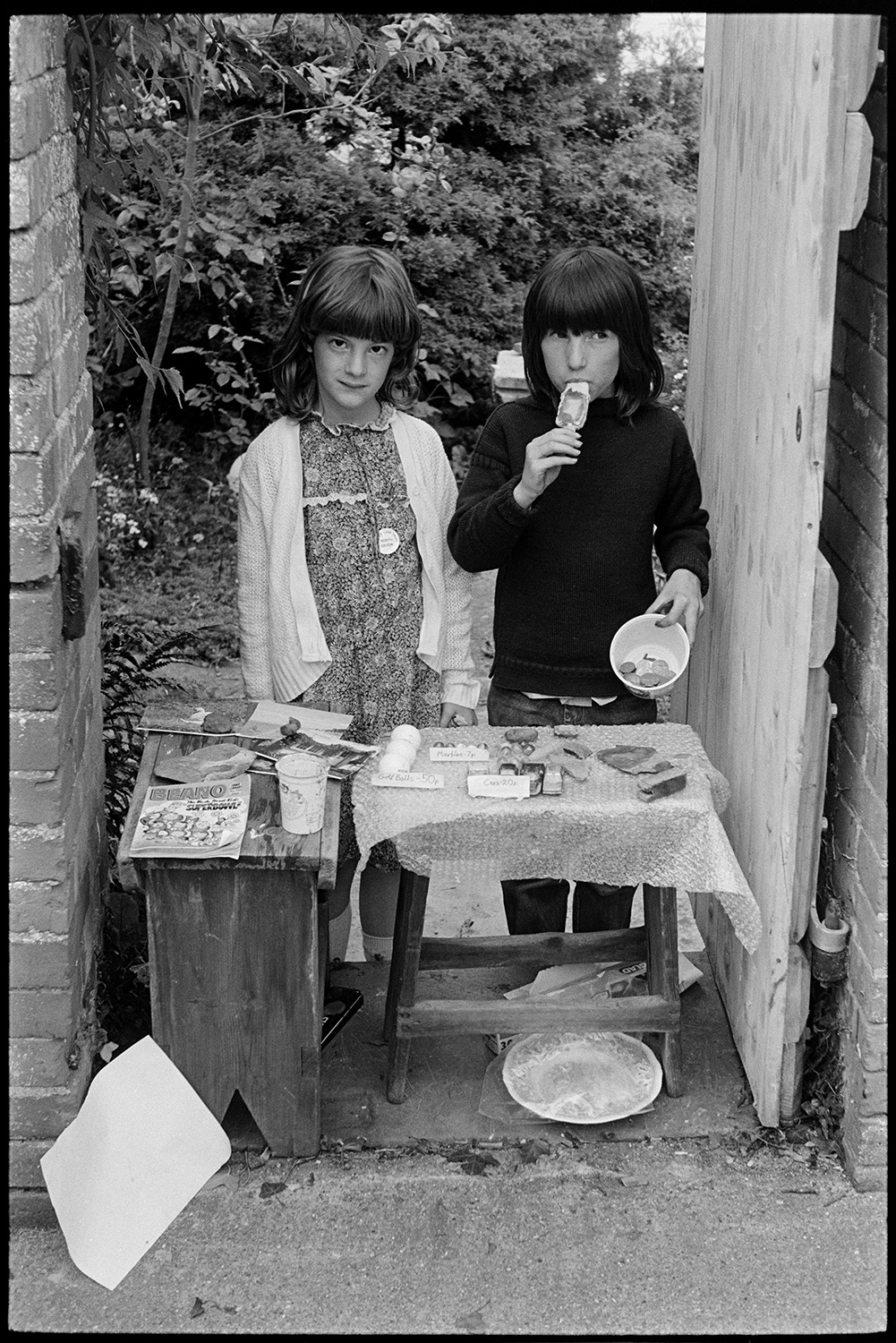 Children's stall on market day of fair.
[Ella Ravilious and Camilla Robinson running a stall by a gate at North Walk, Chulmleigh, on the market day at Chulmleigh Fair. They are selling golf balls, cars and marbles. Photographs and a Beano magazine are also on the stall.]