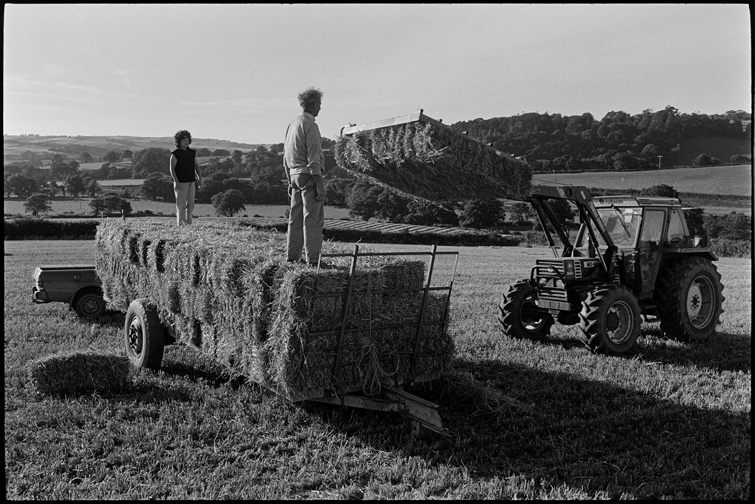 Loading bales of straw.
[A man and woman standing on bales of straw on a trailer while bales are loaded onto the trailer by a tractor, in a field at Rashleigh Barton, Chulmleigh.]