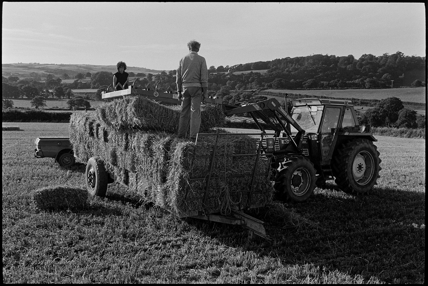 Loading bales of straw.
[A man and woman standing on bales of straw on a trailer while bales are loaded onto the trailer by a tractor, in a field at Rashleigh Barton, Chulmleigh.]