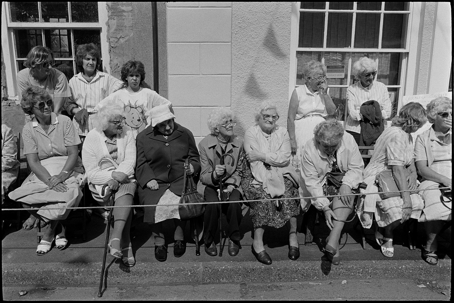 Spectators waiting for start of fair parade.
[Women sitting on seats on the pavement and on windowsills, waiting for parade in Fore Street, Chulmleigh during Chulmleigh Fair.]