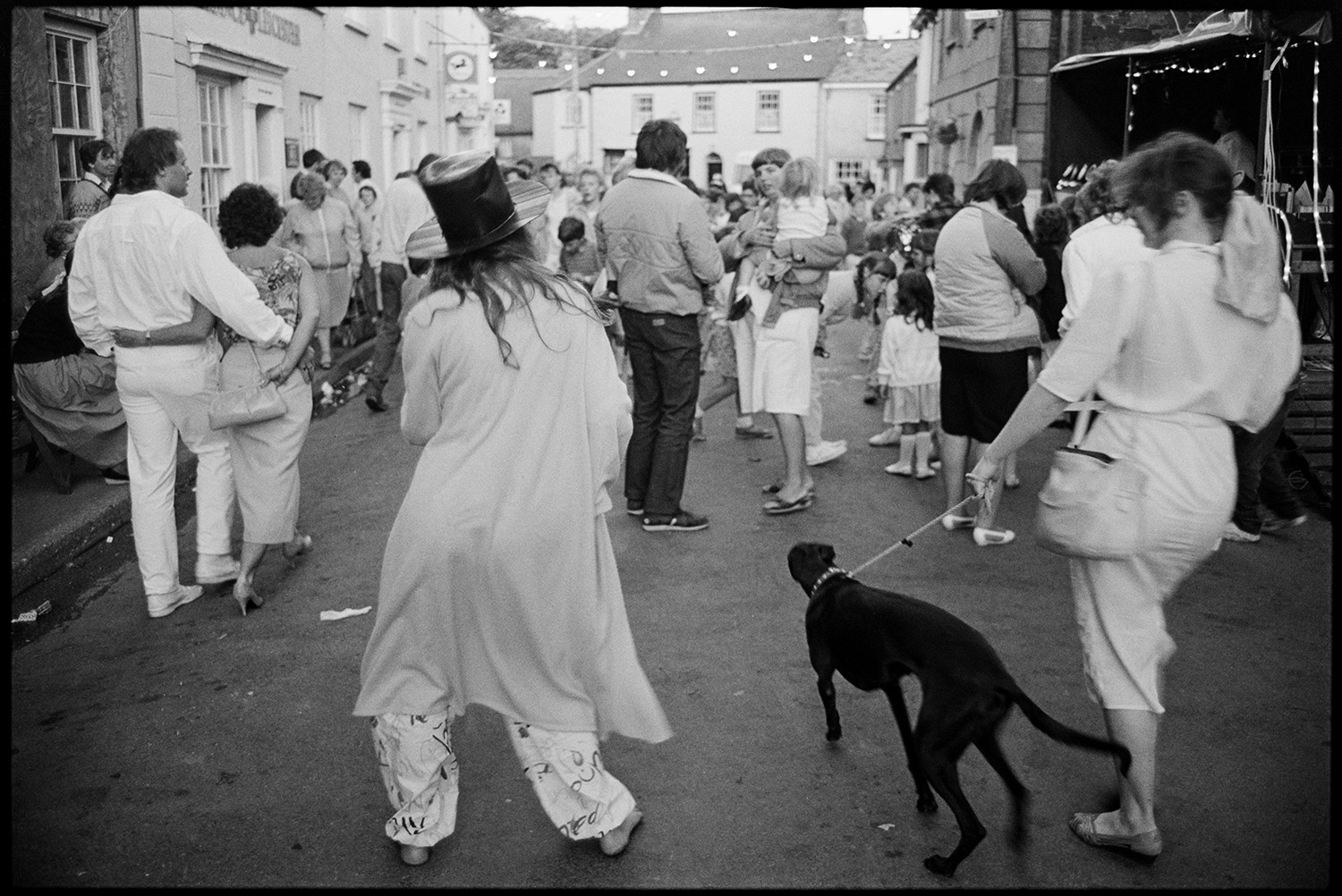 Dancing in the street at fair. Fancy dress.
[People dancing and standing at Chulmleigh Fair in Fore Street, Chulmleigh. The street is decorated with festoon lights.]