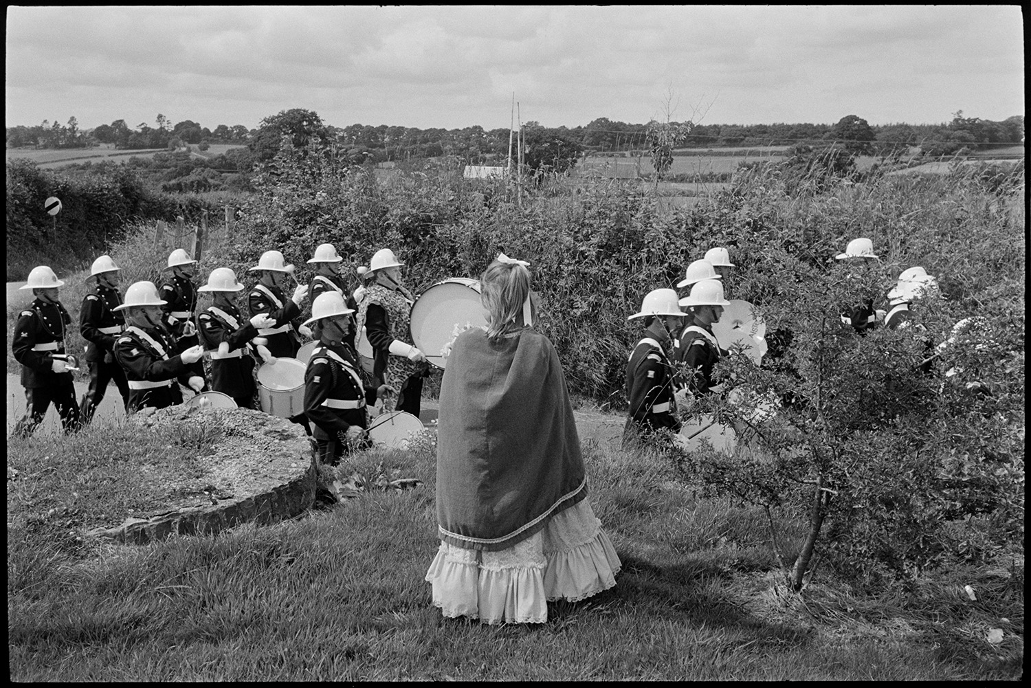 Corps of drums parading before fair procession with queen and fancy dress entries.
[A girl standing in a garden wearing a fancy dress outfit and watching a Corps of drums march by at Chulmleigh Fair.]