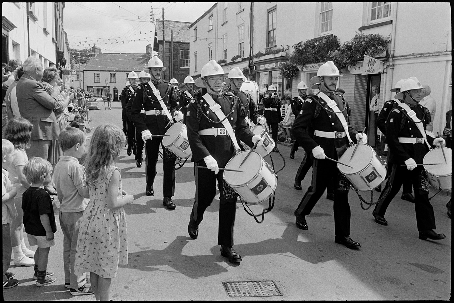 Corps of drums parading before fair procession with queen and fancy dress entries.
[Corps of drums playing and marching down a street with men, women and children lining the street to watch at Chulmleigh Fair. The street is decorated with bunting and shop fronts can be seen in the background.]