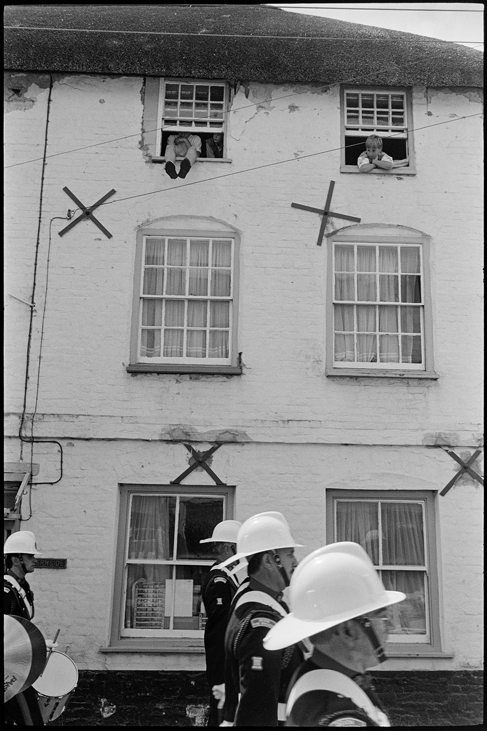 Corps of drums parading before fair procession with queen and fancy dress entries.
[People looking out of upstairs windows at a Corps of drums marching down a street at Chulmleigh Fair.]