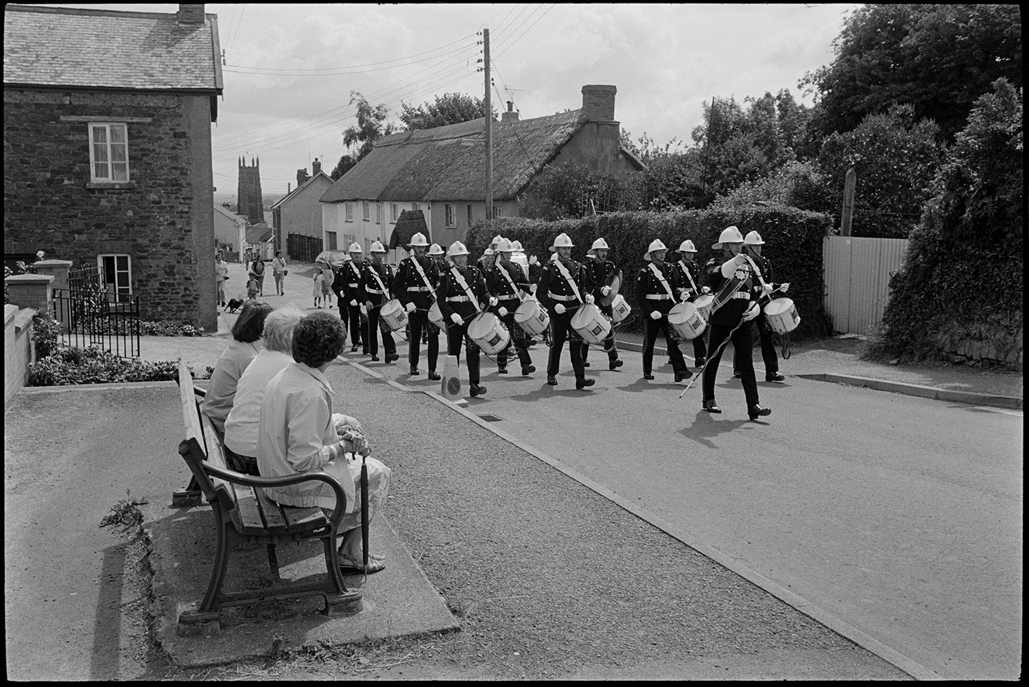 Corps of drums parading before fair procession with queen and fancy dress entries.
[Three women sitting on a bench by a road watching a Corps of drums marching down a street at Chulmleigh Fair. Chulmleigh Church can be seen in the background.]