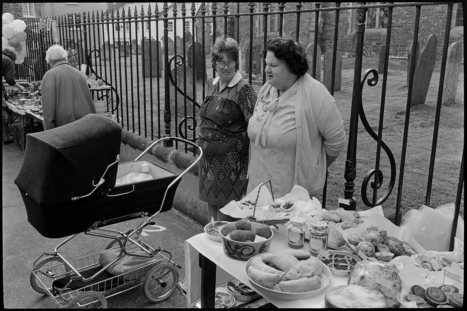Procession, Queen and attendants from church to open fair, money scramble, cake stalls.
[Two women with a pram standing and talking next to a table spread with food by the church railings at Chulmleigh Fair. The churchyard can be seen in the background.]