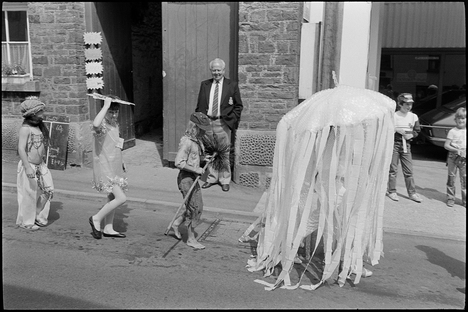 Procession, Queen and attendants parading , fancy dress prizes women spectators laughing !
[Children in fancy dress parading down a street past a man and children watching the Chulmleigh Fair procession. One child is dressed as a jellyfish and another is a chimney sweep.]