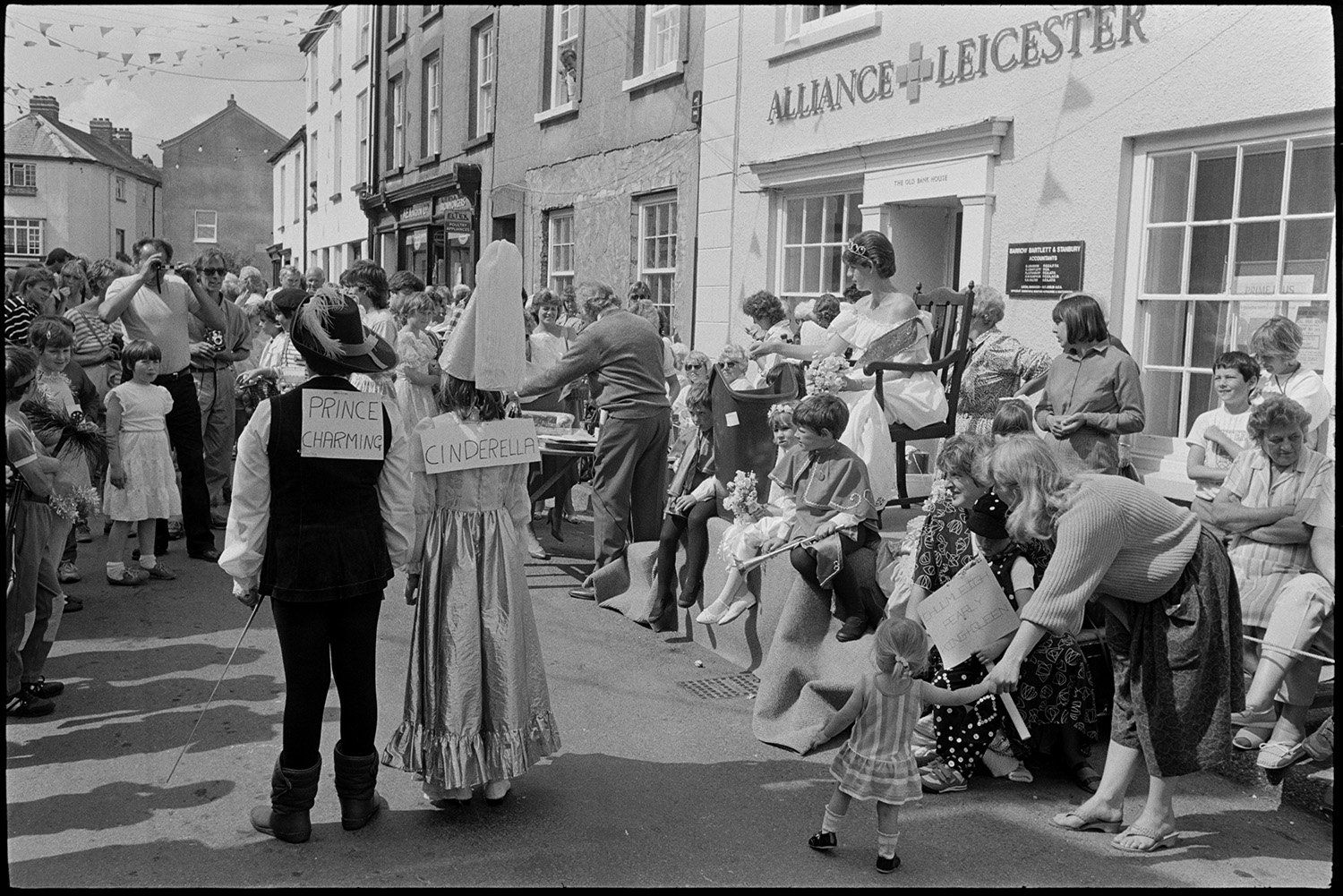 Procession, Queen and attendants parading , fancy dress prizes women spectators laughing !
[People gathered in the street outside the Alliance and Leicester Bank for Chulmleigh Fair,  looking at two children dressed as Prince Charming and Cinderella. A man is taking a photograph of them. Children in fancy dress and Debbie Hawkins, the Chulmleigh Fair Queen, are sitting on a podium and the pavement nearby.]