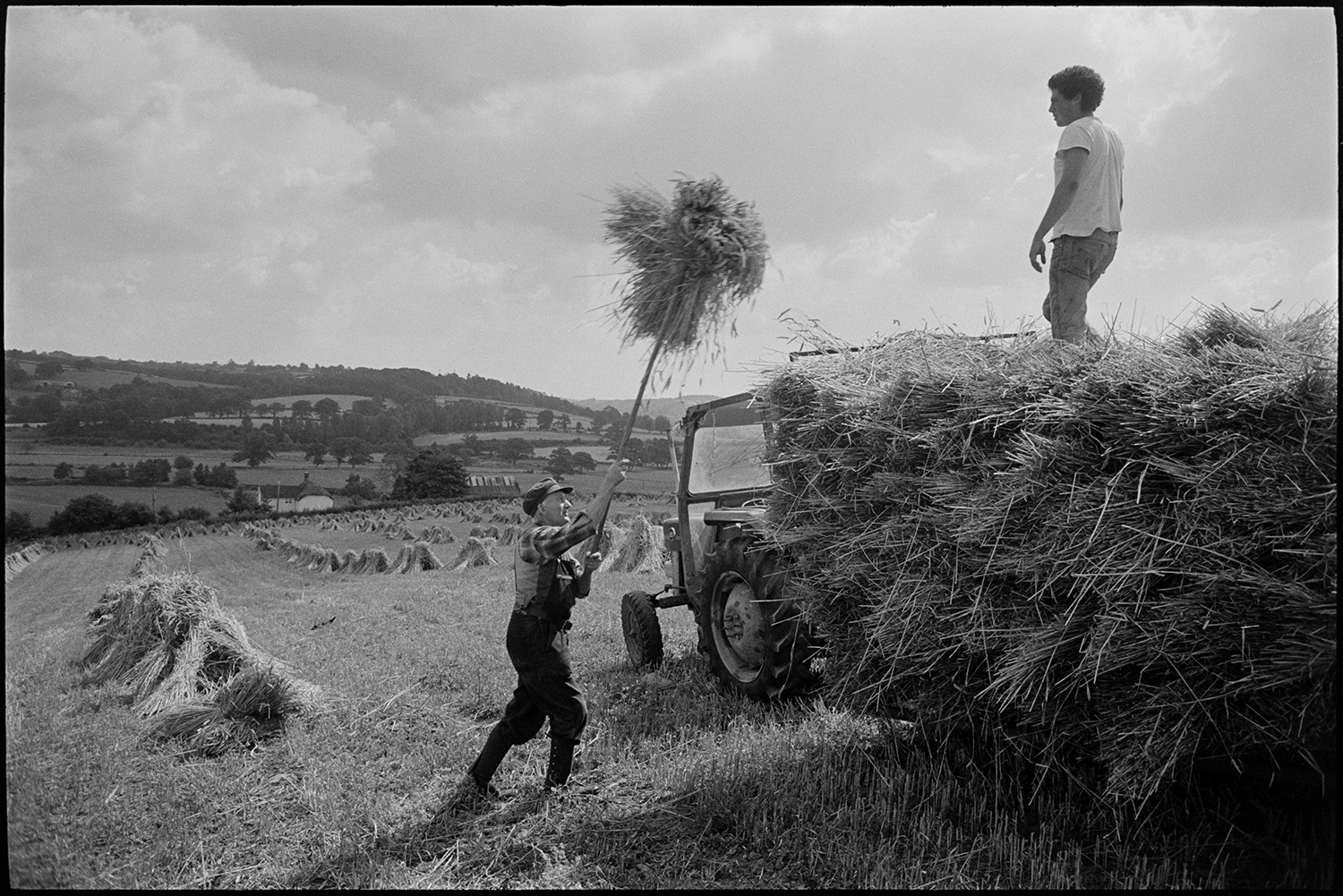 Men building wheatrick in harvest field.
[Men, possibly from the Cole family, loading stooks of corn or wheat onto a trailer using a pitchfork in a field at Ashreigney. Views of hills, fields and trees are visible in the distance.]