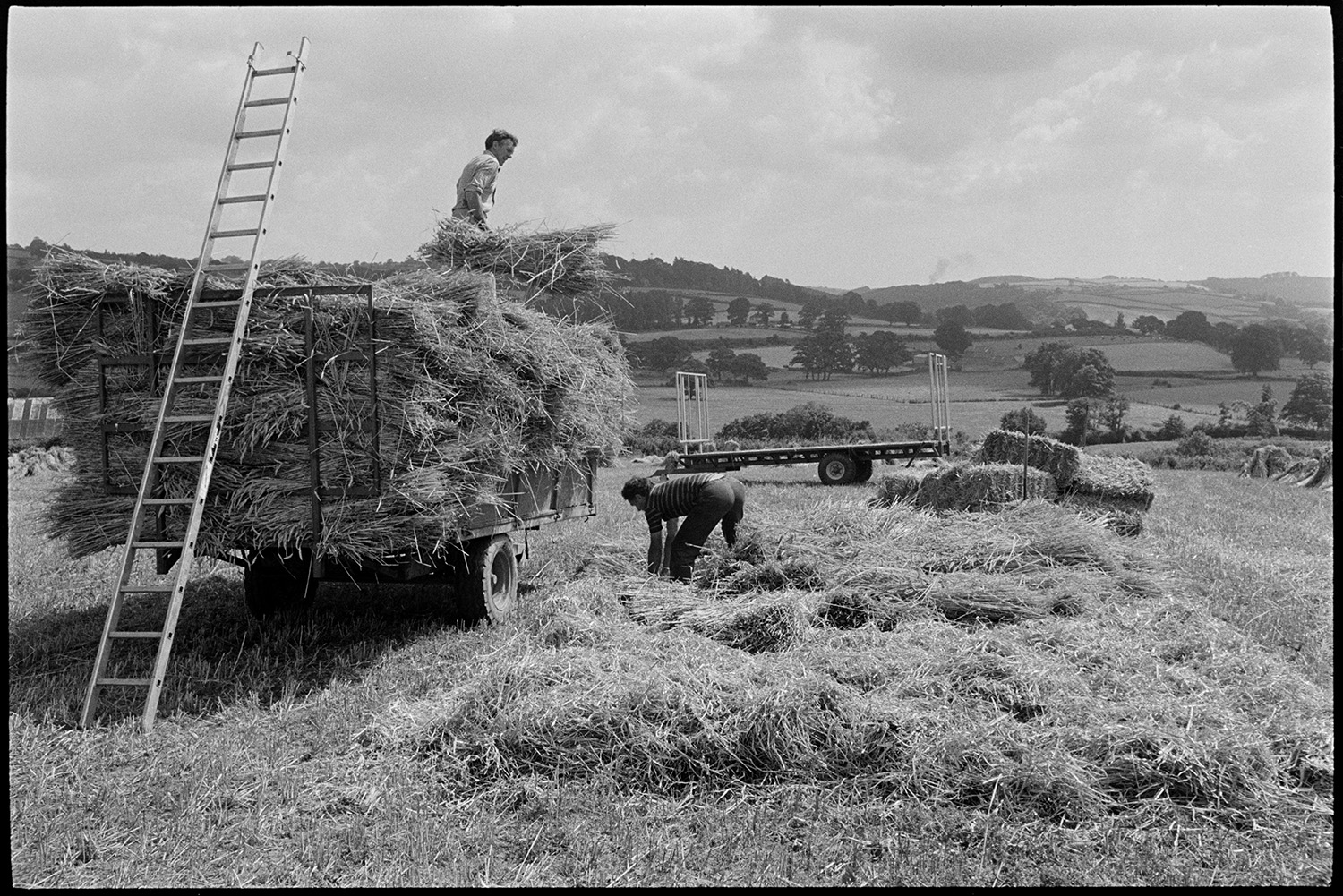 Men building wheatrick in harvest field.
[Men, possibly from the Cole family, unloading  corn or wheat from a trailer and building a wheatrick in a field at Ashreigney. A ladder is resting against the trailer. Views of hills, fields and trees are visible in the distance.]