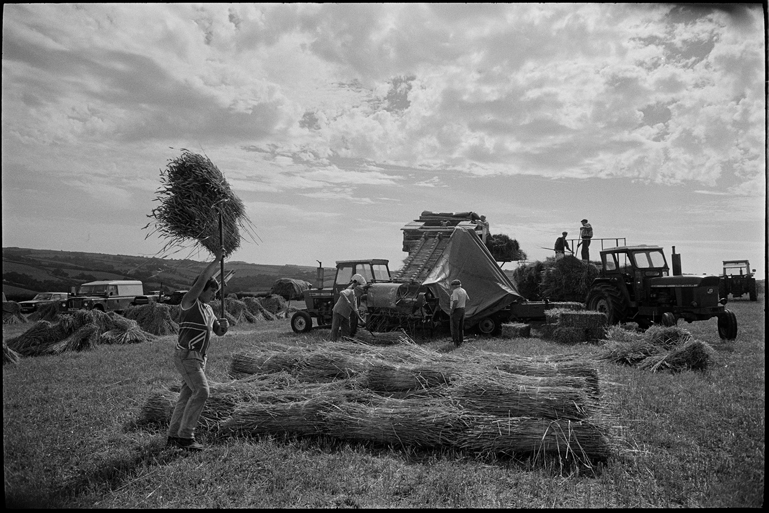 Reedcombing in harvest field men carrying nitches of wheat to reed comber.
[A group of men and a boy working with a reed comber in a field at Spittle, Chulmleigh. Tractors and a Land Rover are parked in the field. Bryan Down is the boy in the foreground wearing a Union Jack t-shirt. He is carrying bundles of reed above his head with a pitchfork to add to a pile of reed in the foreground.]