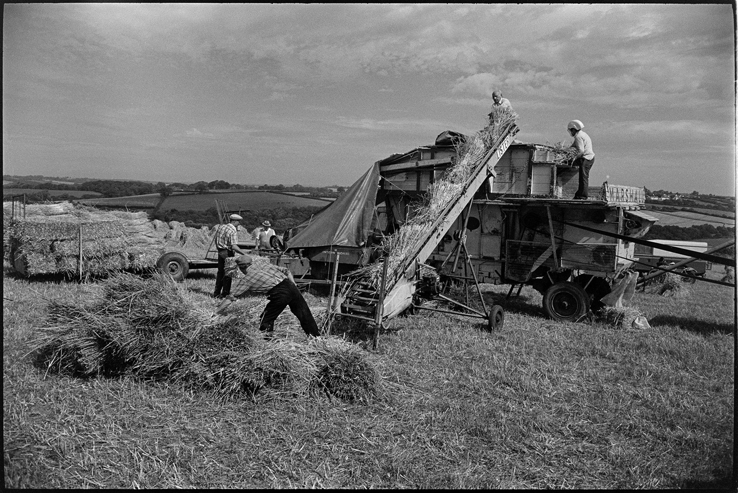 Reed combing in harvest field men carrying nitches of wheat to reed comber.
[Men, possibly from the Down family, working with a reed comber in a field at Spittle, Chulmleigh. One man is  lifting bundles of wheat onto the elevator to be processed by the reed comber. Another man on the reed comber is wearing a helmet.]
