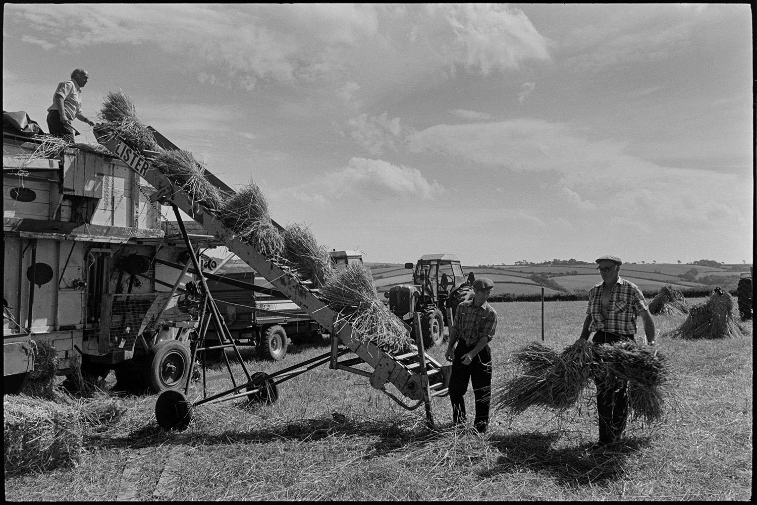 Reed combing in harvest field men carrying nitches of wheat to reedcomber.
[Men, possibly from the Down family, working with a reed comber in a field at Spittle, Chulmleigh. One man is passing bundles of wheat to another man who is lifting them onto the elevator to be processed by the reed comber.]