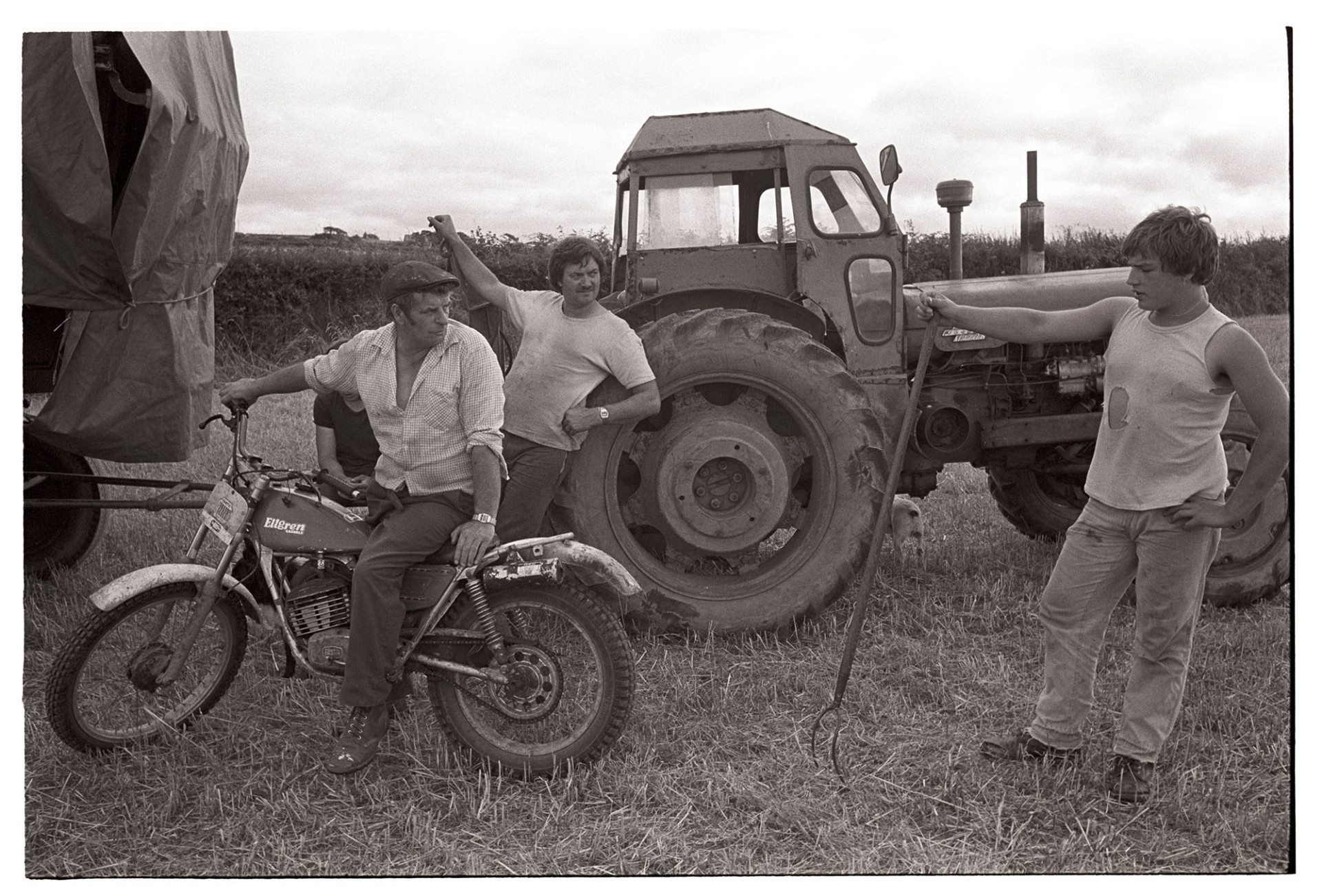 Farmer on motorbike talking to harvesters. 
[Mr Down on a motorbike talking to two harvesters in a field at Spittle, Chulmleigh. One of the men is leaning on a pitchfork. They are stood by a tractor and trailer. The trailer is covered with polythene.]