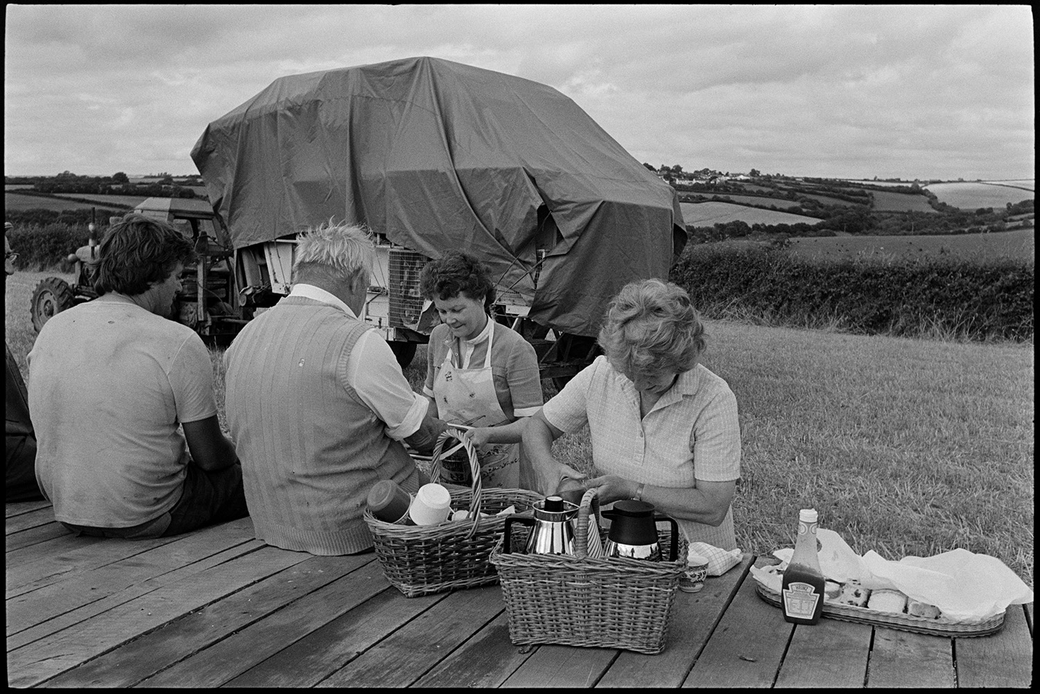 Woman farmers wife serving refreshments to harvesters in the field. 
[Pam Down, on the left, and another woman serving refreshments to men reedcombing in a field at Spittle, Chulmleigh. The men are sat on a trailer. The reed comber can be seen in the background under a polythene sheet.]