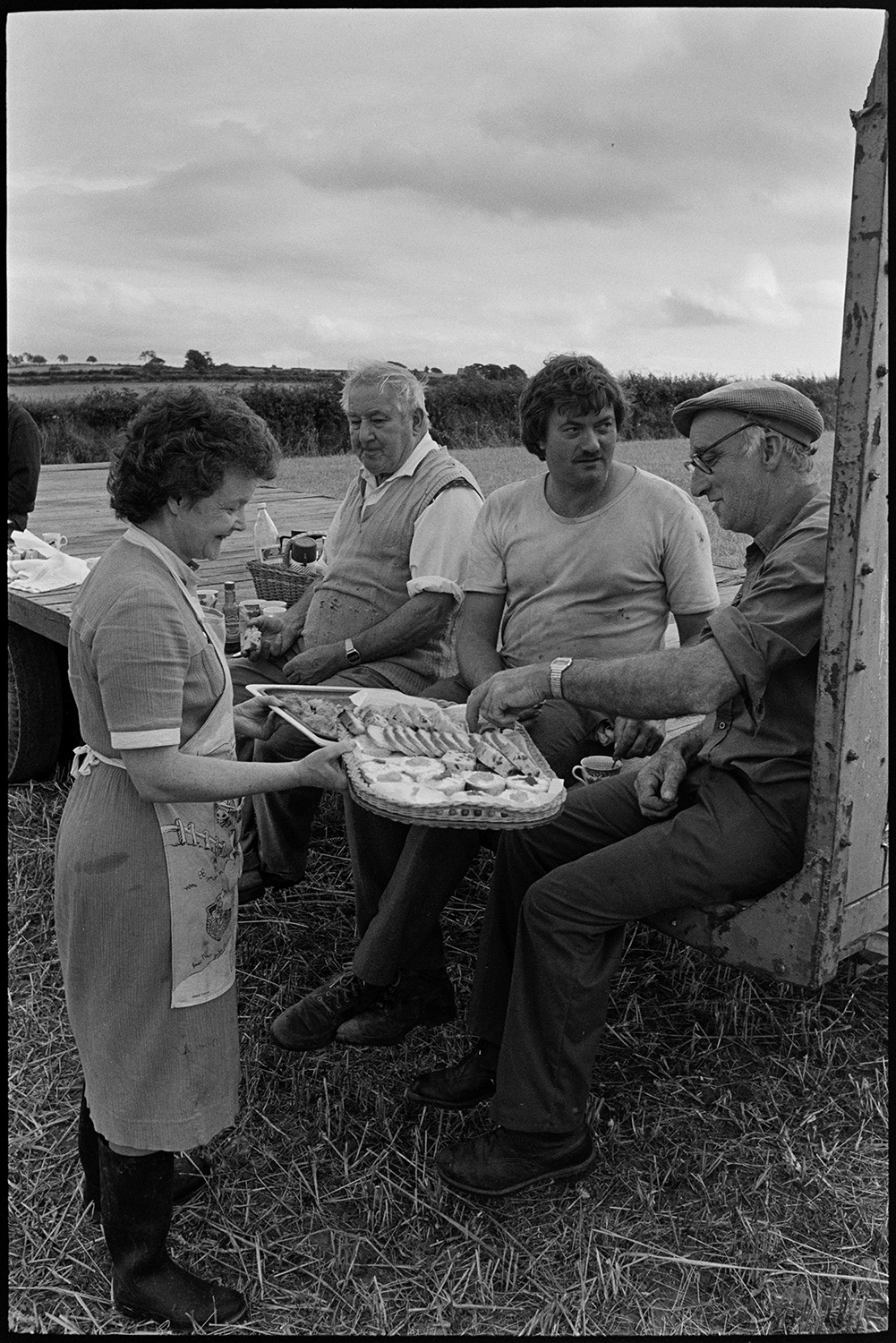 Woman farmers wife serving refreshments to harvesters in the field. 
[Pam Down serving slices of cake on trays to men reedcombing in a field at Spittle, Chulmleigh. The men are sat on a trailer.]
