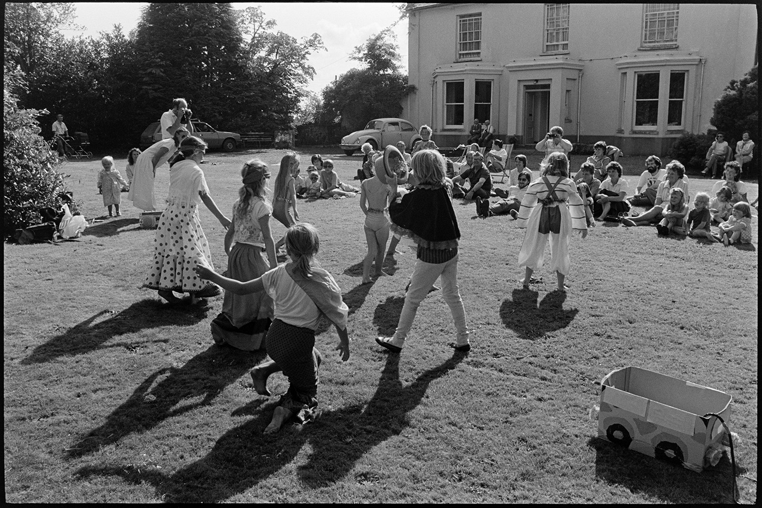 Children's ballet class dance performance on lawn of country house. 
[Children performing a ballet routine to their parents, other adults and children on the lawn in front of Broadwoodkelly Rectory, which can be seen in the background.]