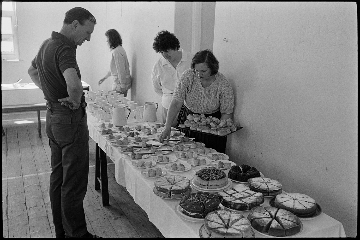 A man looking at a refreshment table at a children's ballet class in Broadwoodkelly Village Hall. A woman is putting out scones onto plates.