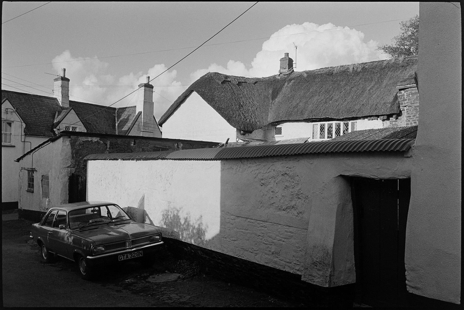Street scenes, cottages, pub and chemist, mothers and children. 
[A car parked by a wall with a corrugated iron cap in a street in Chulmleigh. A thatched cottages can be seen behind the wall.]