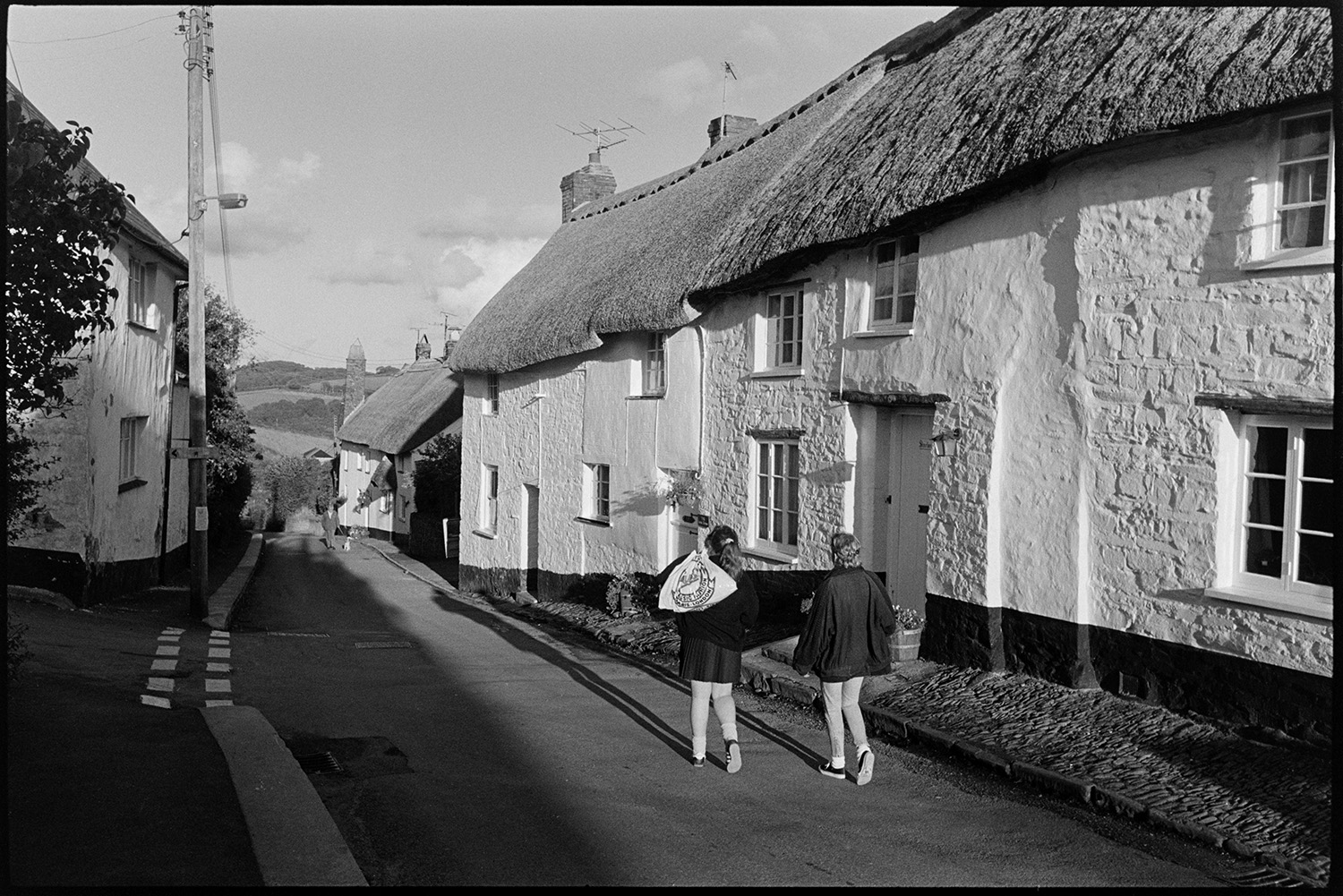 Street scenes, cottages, pub and chemist, mothers and children. 
[Two children walking down a street in Chulmleigh, past thatched cottages. A man and dog are walking up the street towards them.]