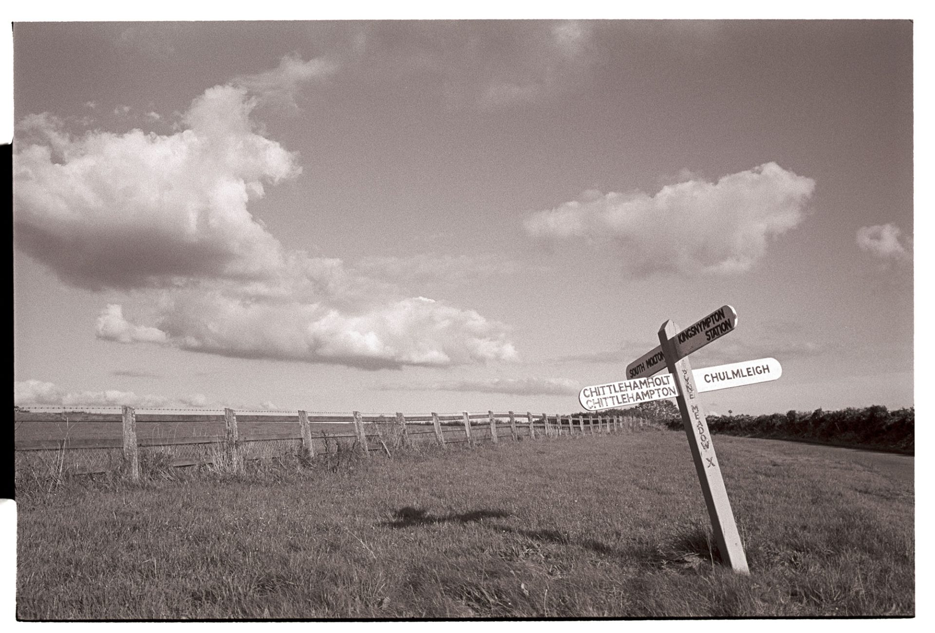 Road sign at lonely crossroads. 
[A signpost by a field at Pyne Meadow Cross, Chulmleigh. The signpost is listing to one side.]