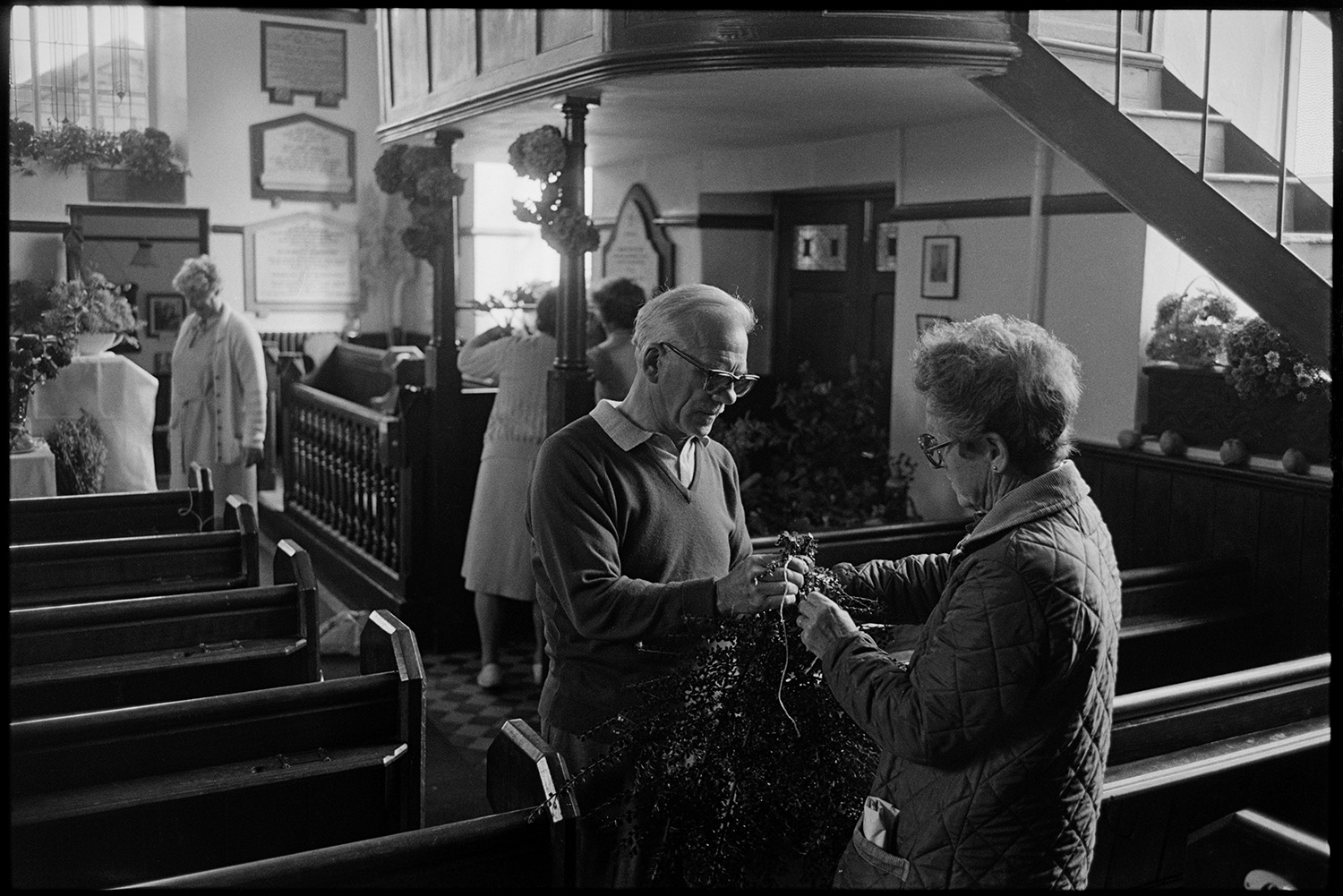Congregational chapel interior with people decorating for Harvest Festival and having tea. 
[A man and woman tying together foliage for a display for the Harvest Festival in Chulmleigh Congregational Church. Pews and steps leading to the gallery can be seen inside the church.]