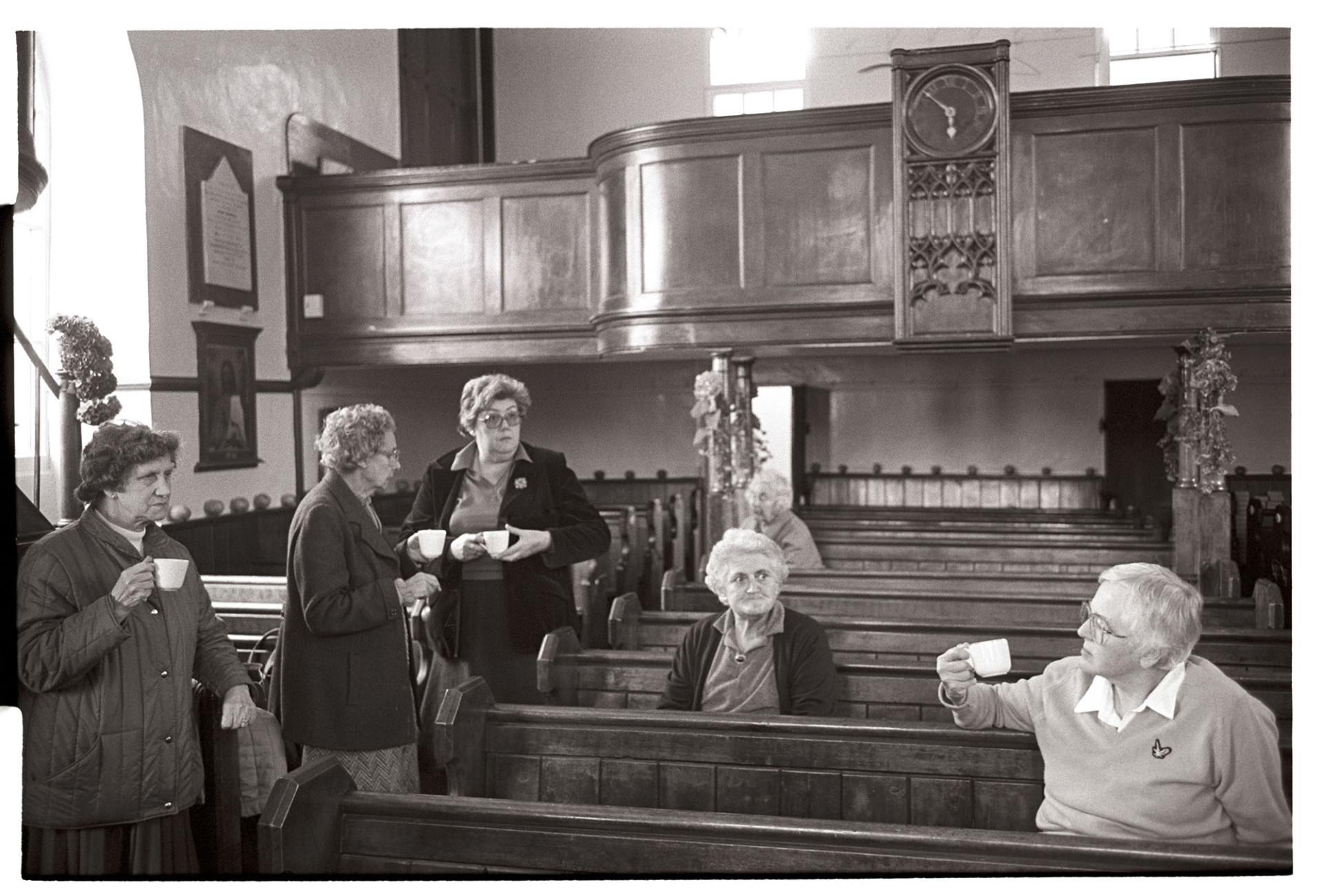 Congregational chapel interior tea or coffee break for people decorating at Harvest Festival. 
[Women, including Elaine Marsh the Minister, having a tea break after decorating Chulmleigh Congregational Chapel for the harvest festival. They are sat in pews and a gallery or balcony with a clock can be seen in the background.]
