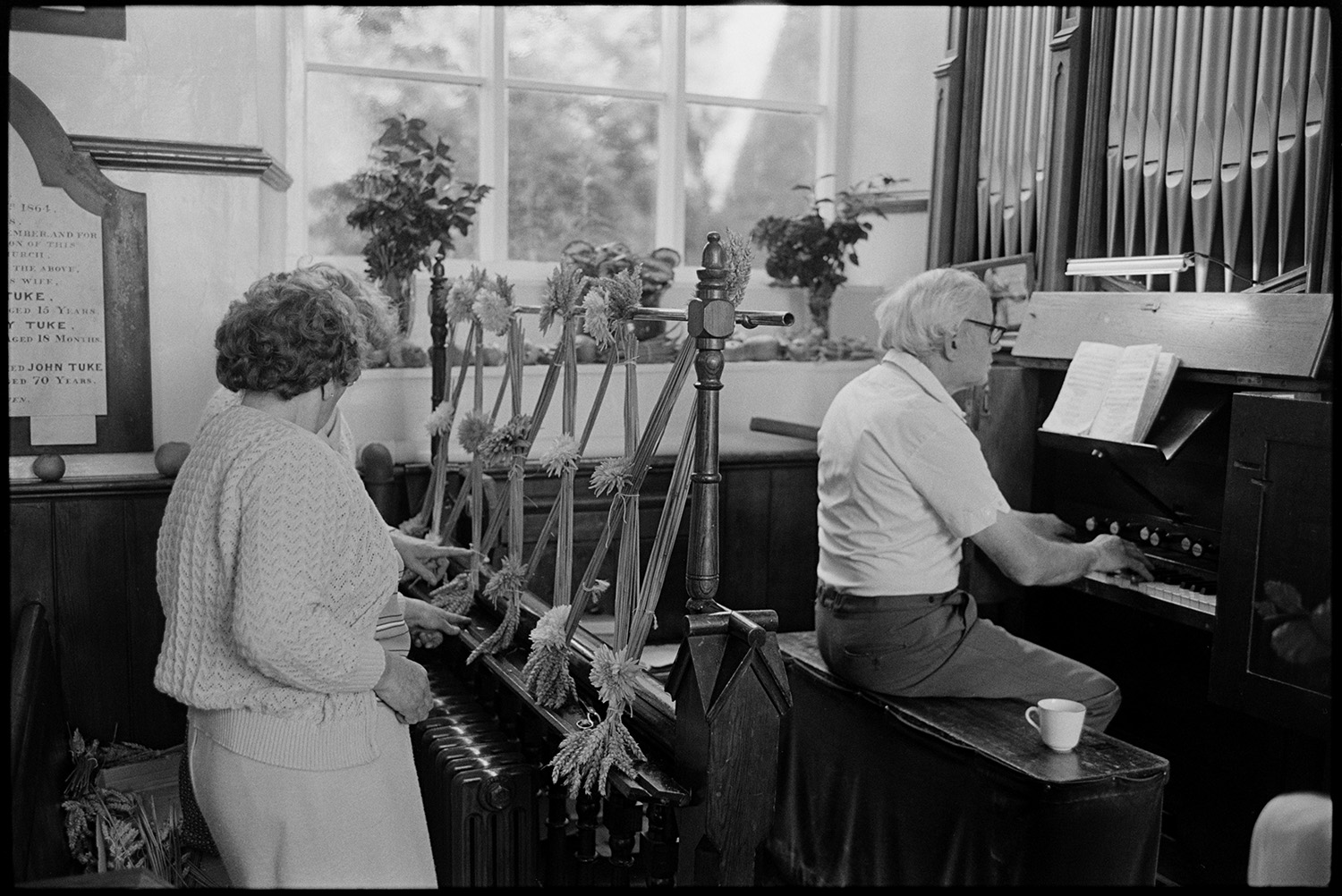Congregational chapel interior with people decorating for Harvest Festival and having tea. 
[Two women arranging a floral display with sheaves of corn for the Harvest Festival by a man playing the organ at Chulmleigh Congregational Chapel. One of women may be Elaine March, the Minister]