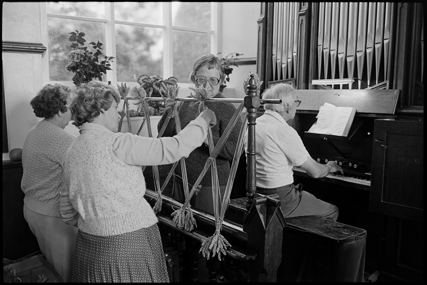 Congregational chapel interior, man playing organ to people decorating at Harvest Festival.
[A man playing the organ at Chulmleigh Congregational chapel while three woman decorate the chapel behind him for the Harvest Festival using tied reeds of wheat. The metal organ pipes are shown above the organ.]