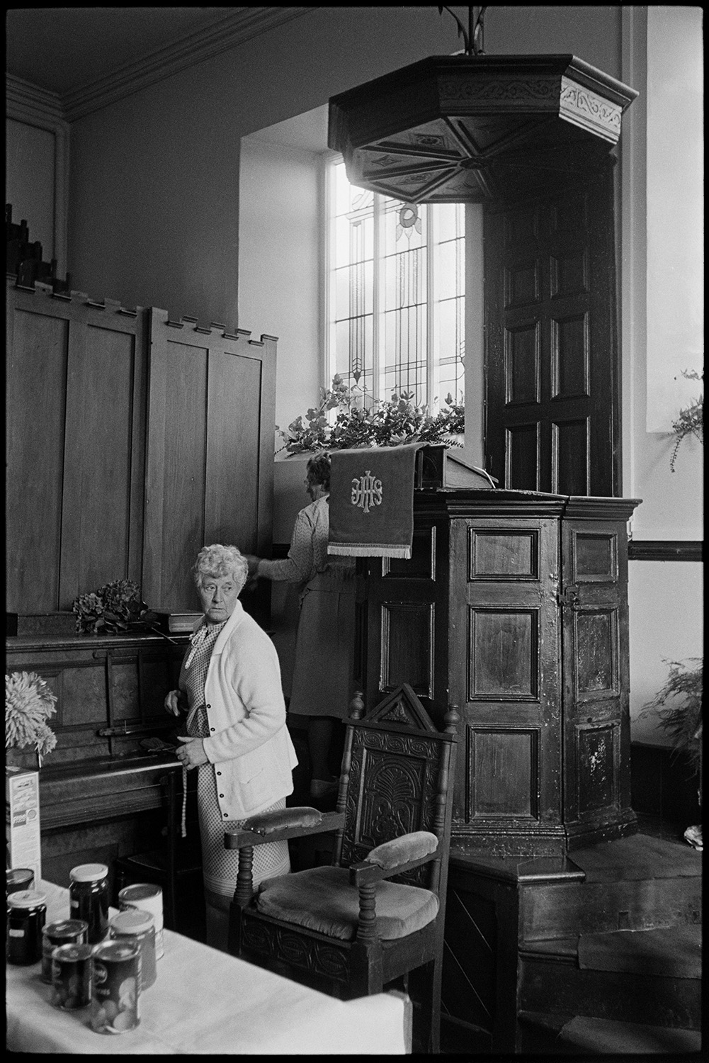 Congregational chapel interior, man playing organ to people decorating at Harvest Festival.
[Two women in Chulmleigh Congregational chapel helping with preparation for the Harvest Festival. An ornate wooden chair is in front of a table with tinned goods and jars for harvest. A wooden screen, pulpit and canopy alongside a decorated window are also visible.]