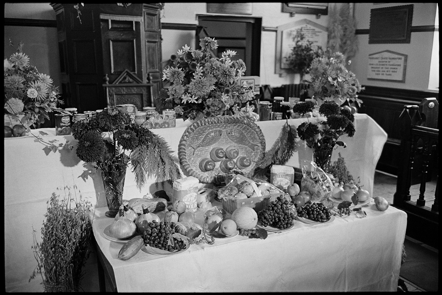 Congregational chapel interior, produce, loaf, on alter table and decorating at Harvest Festival.
[Display of produce at the Harvest Festival in Chulmleigh Congregational chapel. There are vases of flowers, canned food and jars of produce, plates of fruit and vegetables and a salt dough loaf of bread decorated with fives loaves and two fish. A pulpit, chair, memorial wall tablets, and a wooden pew are in the background.]