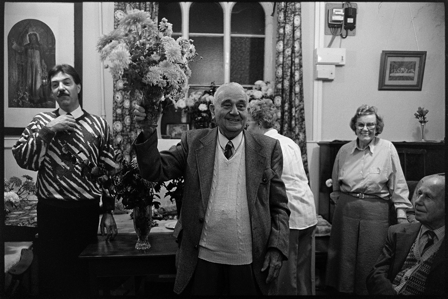 Congregational chapel Harvest supper, sale of produce after supper.
[Sale of produce after the Harvest Festival supper at Chulmleigh Congregational chapel. A man is holding up a bunch of flowers. A window and table with goods and vases of flowers is in the background, with other people watching.]