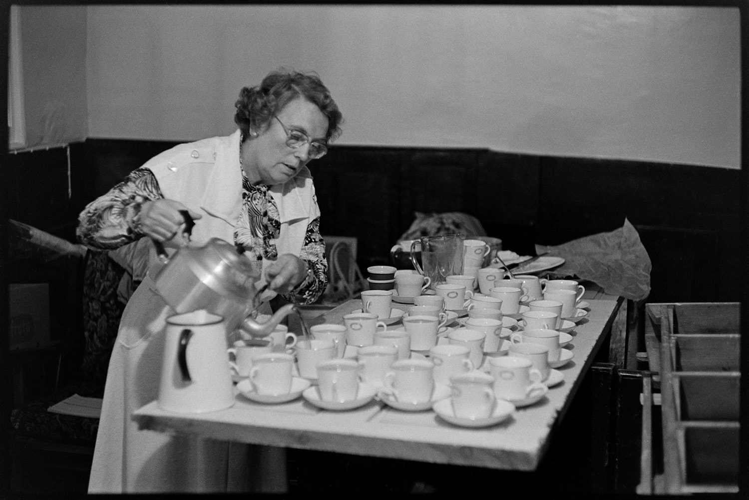Congregational chapel Harvest supper, sale of produce after supper. Women chatting.
[A woman pouring tea after the Harvest supper in Chulmleigh Congregational chapel. White china cups and saucers and a white enamel jug are laid out on a wooden table.]