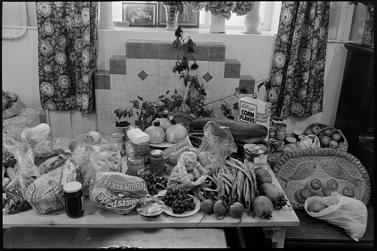 Congregational chapel Harvest supper, singing after meal, sale produce on display.
[A display of produce for sale at the Harvest Festival supper in Chulmleigh Congregational chapel. There is canned food, jars of produce, cereals, fruit, vegetables and a salt dough loaf of bread decorated with fives loaves and two fish. A curtained window with vases of flowers and a tiled fireplace is in the background.]