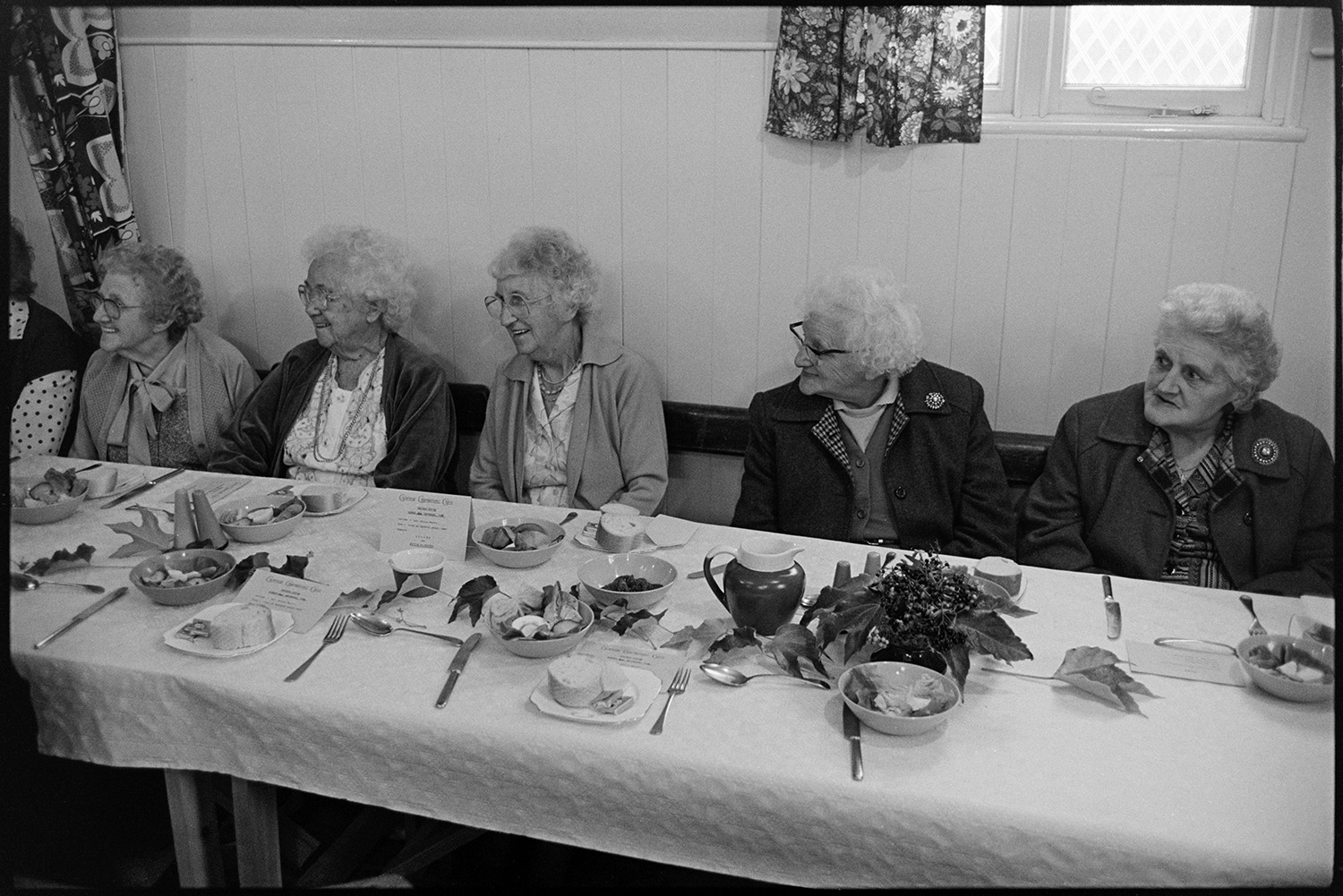 Congregational chapel Harvest supper, singing after meal, sale produce on display.
[Five elderly women sitting at a table at the Chulmleigh Congregational chapel Harvest supper. There is cutlery, crockery, table decorations and plates of bread rolls laid out on the table in front of them.]