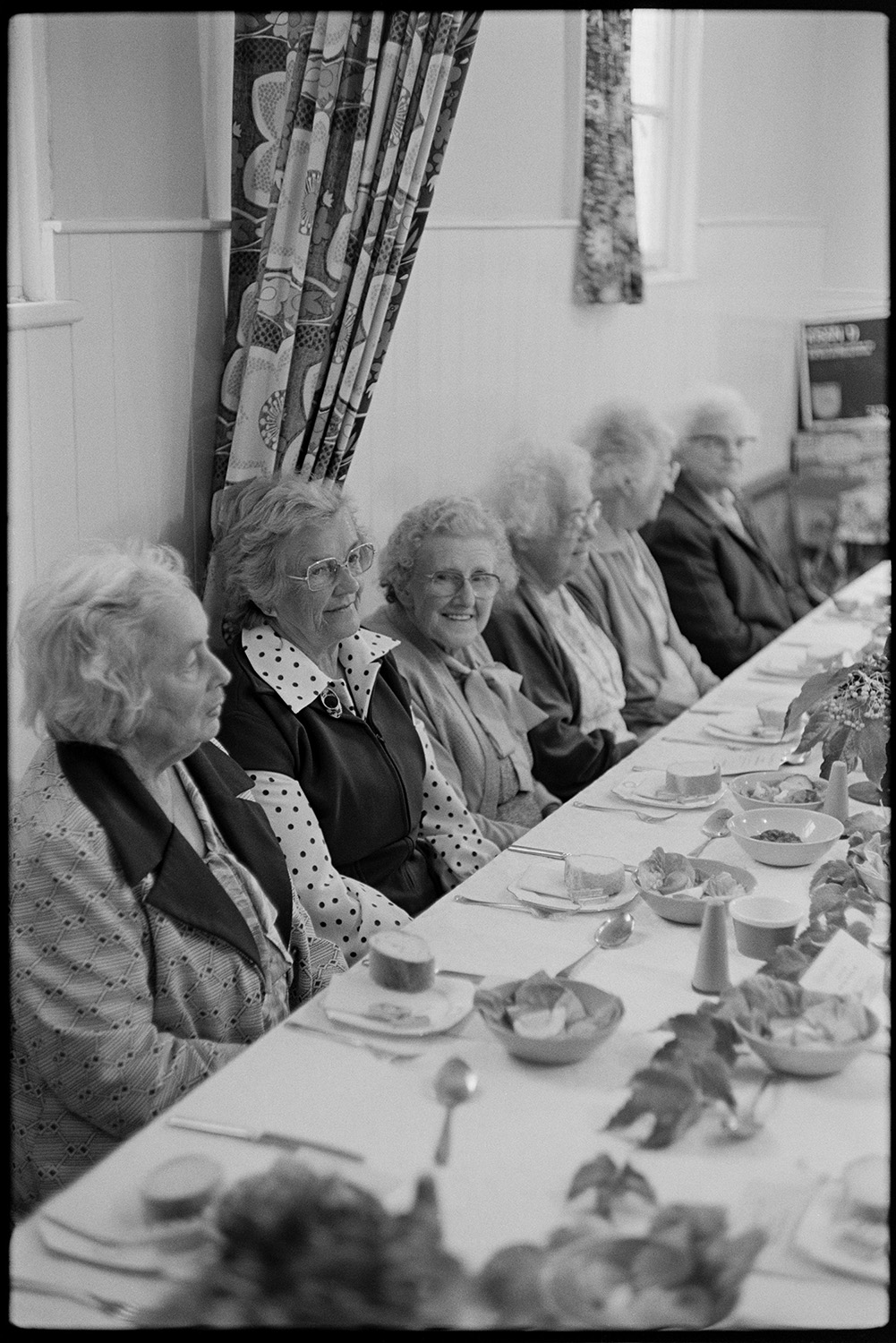 Congregational chapel Harvest supper, singing after meal, sale produce on display.
[Six elderly women sitting at a table at the Chulmleigh Congregational chapel Harvest supper. There is cutlery, crockery, table decorations and plates of bread rolls laid out on the table in front of them.]