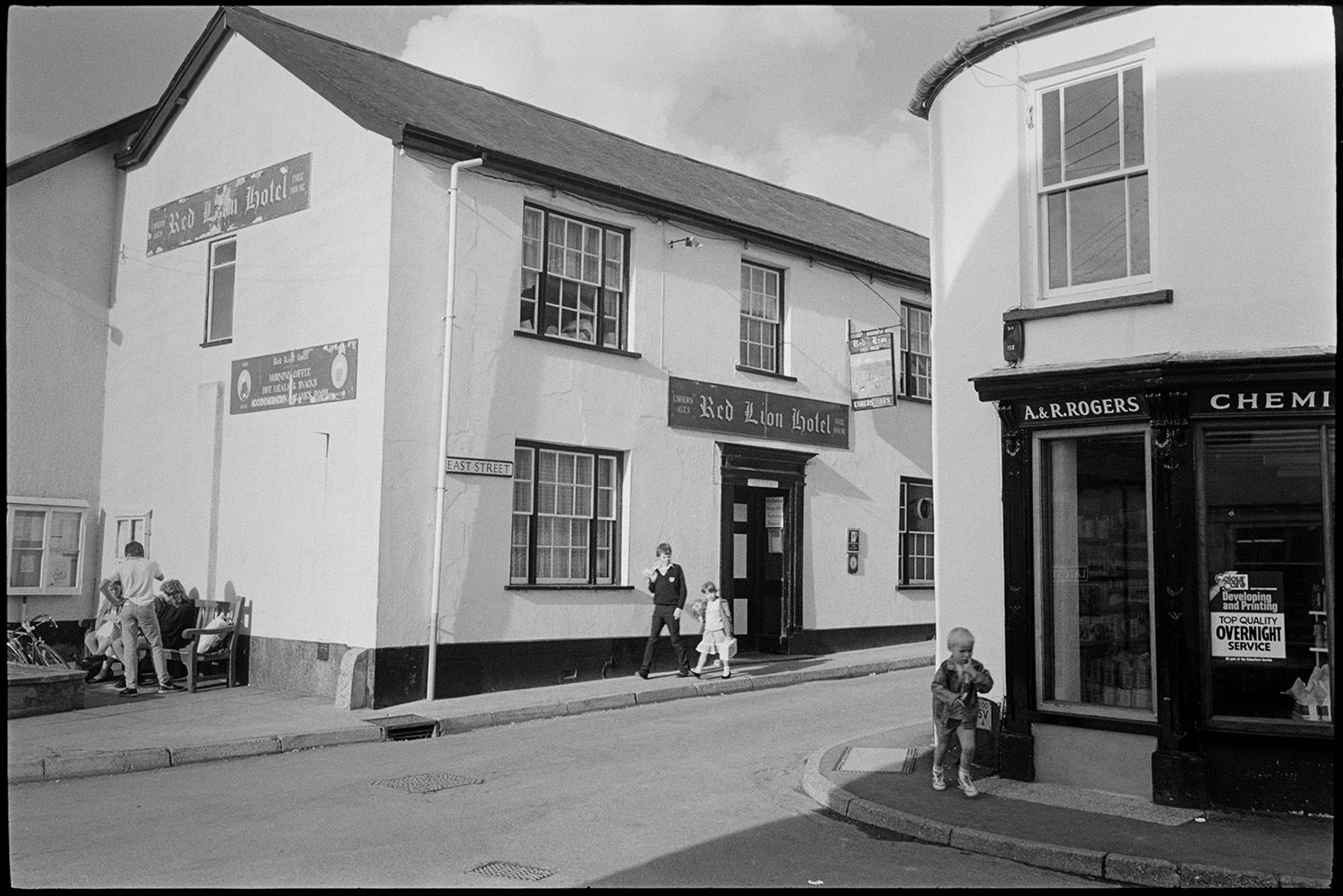Street scenes with children practising throwing a baton.
[The corner of East Street in Chulmleigh, showing the Red Lion Hotel on one side and A & R Rogers the Chemists on the other. There is a noticeboard and a wooden bench with people sitting on it by the hotel and young children are walking along the pavement.]