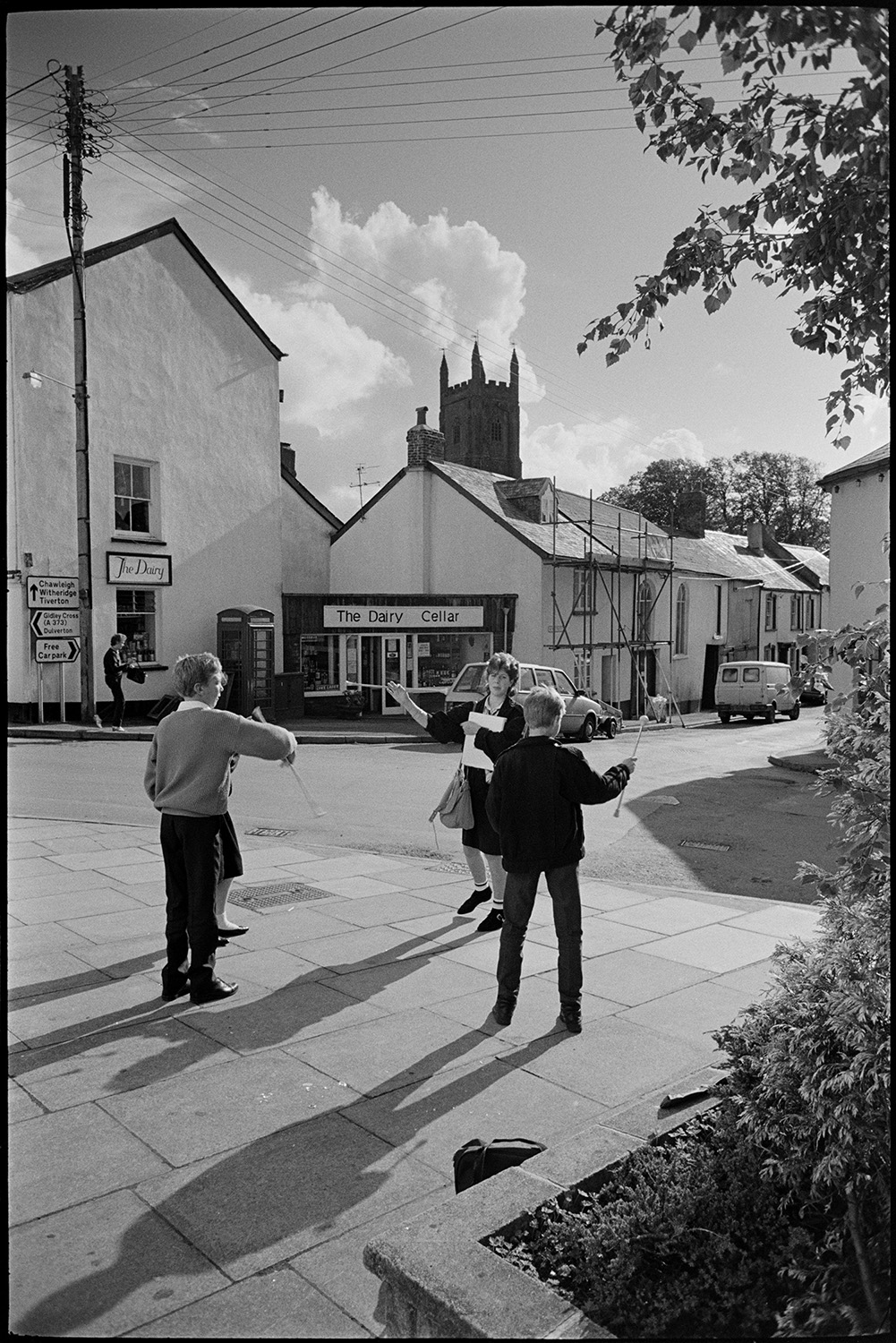 Street scenes with children practising throwing a baton.
[Children practising throwing a baton on a street corner in Chulmleigh. Chulmleigh church tower is in the background with town houses, a telephone kiosk, and the shop front of The Dairy Cellar.]