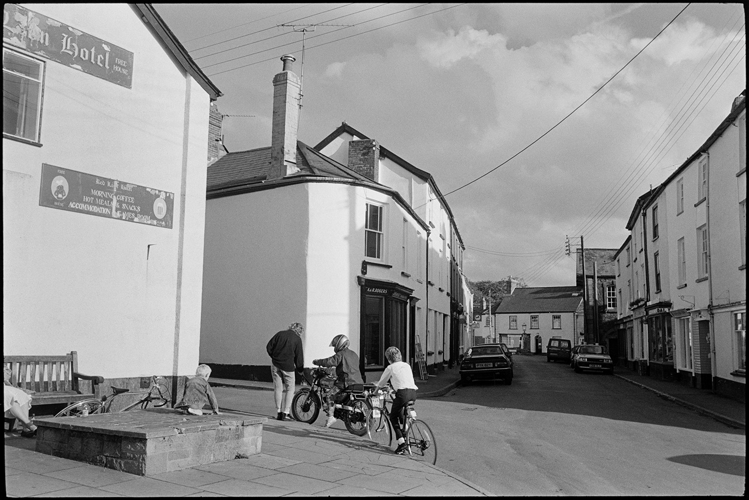 Street scenes with children practising throwing a baton.
[A street scene in Chulmleigh showing the Red Lion Hotel and A & R Rogers the chemists, with the main street, shops and town square in the background. Young people with a motorcycle and bicycles are passing by a wooden bench on the pavement in the foreground.]