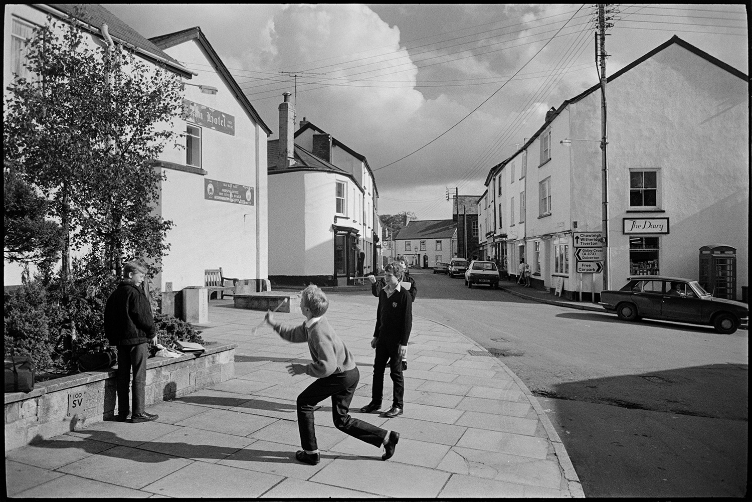 Street scenes with children practising throwing a baton.
[Children practising throwing a baton on a street corner in Chulmleigh. The Red Lion Hotel is in the background with town houses, a telephone kiosk, and The Dairy shop front.]