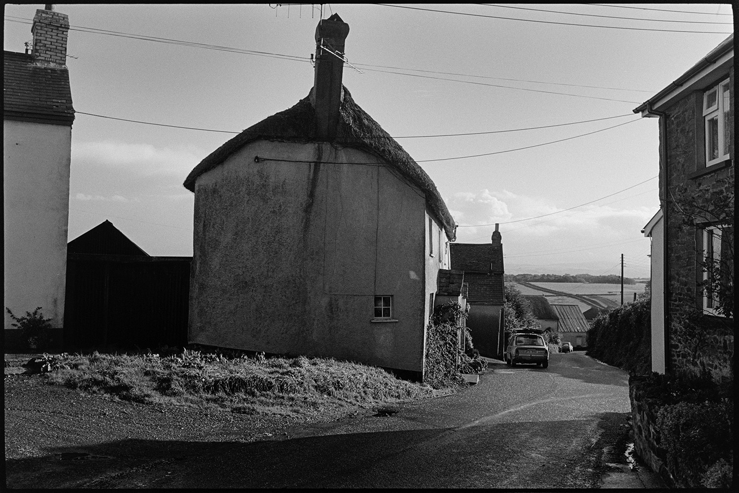 Road with thatched cottage at lower end of village.
[A road at the lower end of Winkleigh, showing a thatched cottage, other village houses and hills and fields in the distance.]
